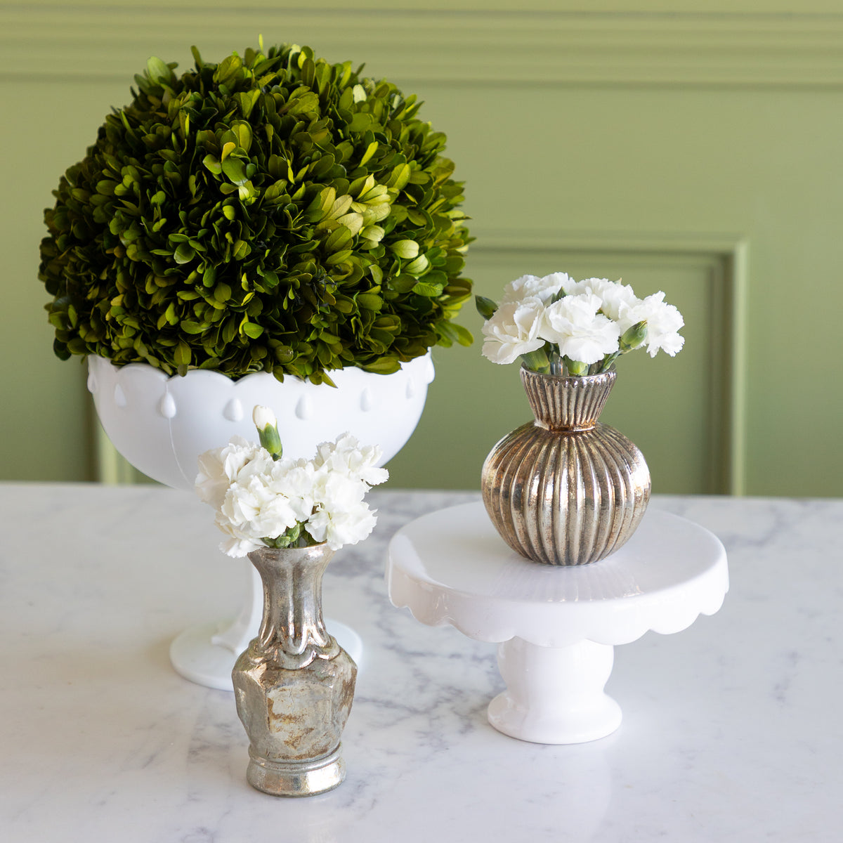 Three decorative vases and floral arrangements displayed on a white marble surface against a sage green paneled wall. The arrangement includes a large round green boxwood top in a white pedestal bowl, a small white flower cluster in a ribbed metallic gold vase, and another white flower cluster in a small silver vase.