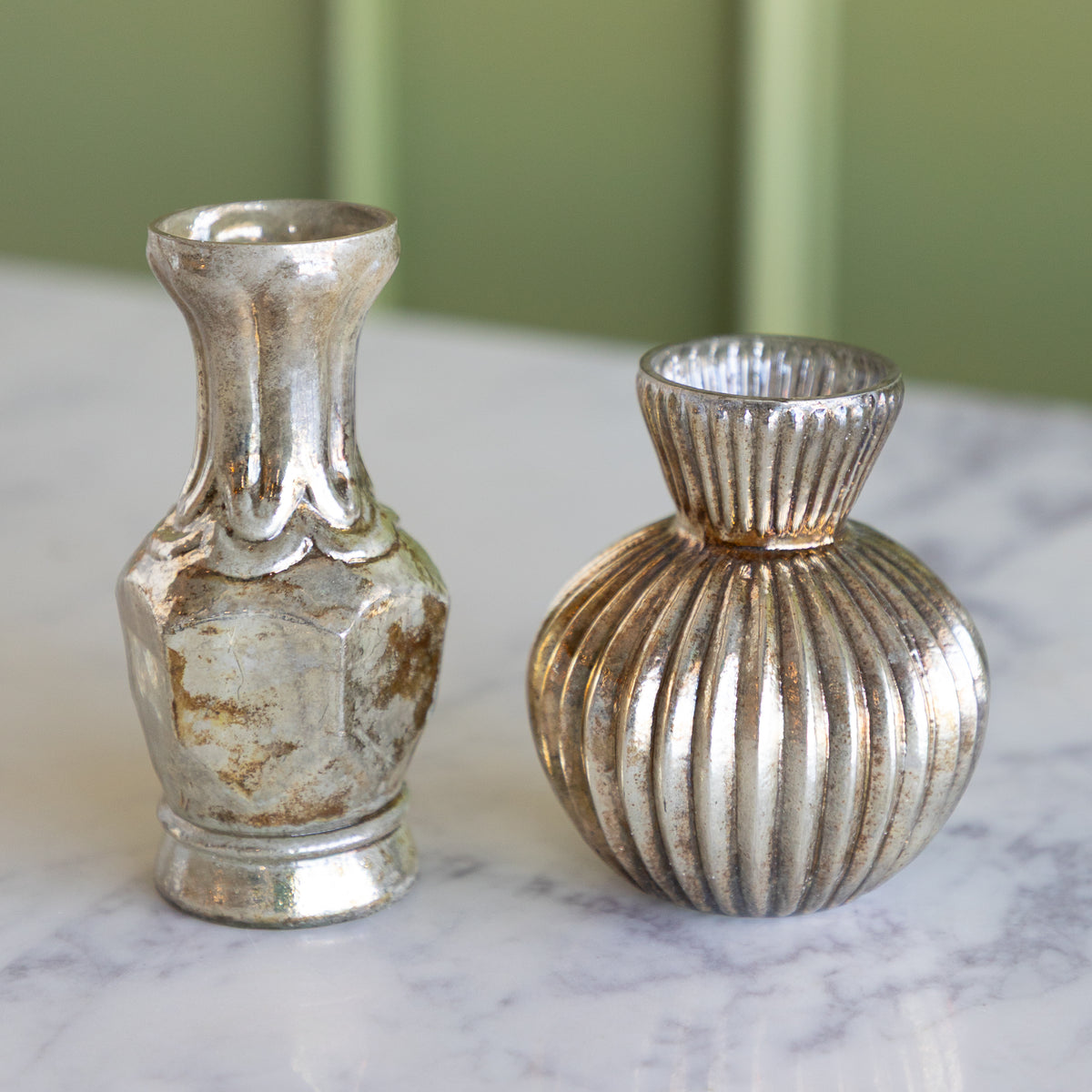 Two decorative silver-toned metal vases with distressed finishes, one with a flared neck and one ribbed round vase, displayed on a white marble surface against a soft green background.