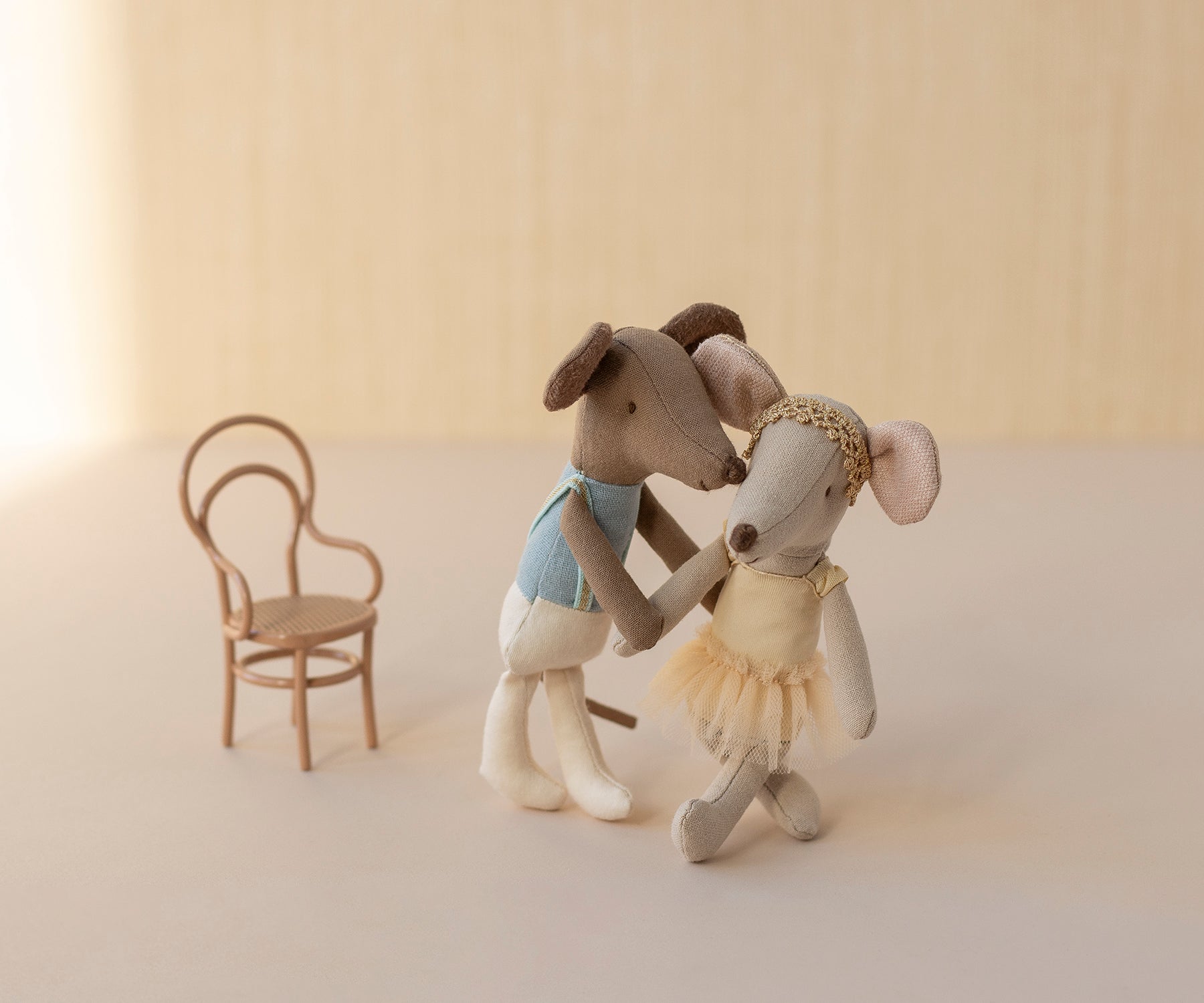 Two stuffed animal toys, one in a dress and one in a suit, standing next to a small chair on a light background.