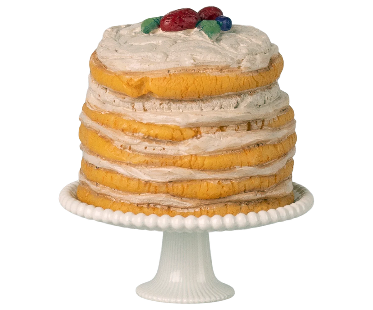 A miniature toy layered cake with white frosting and fruit on top, placed on a white cake stand.