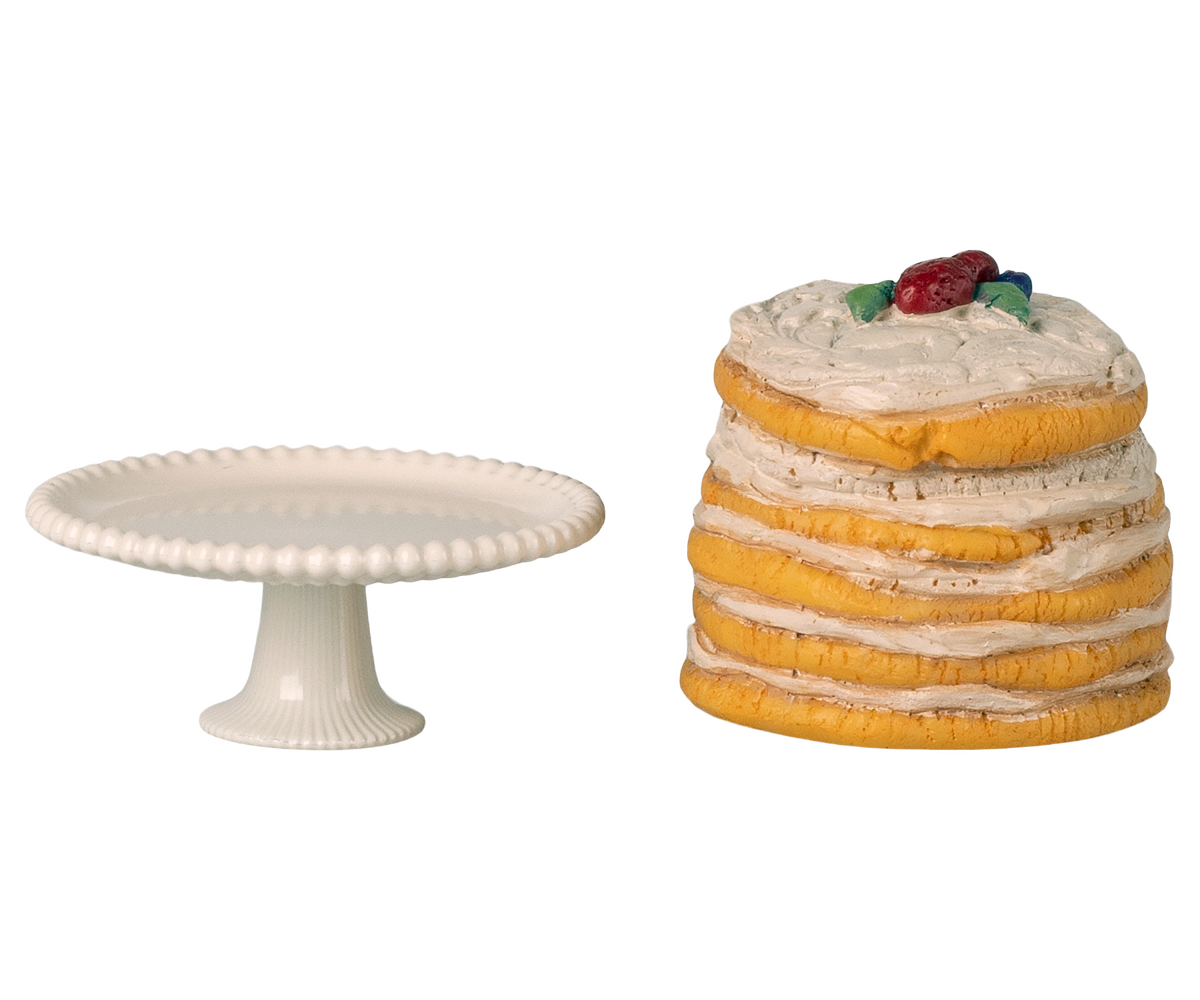 Stack of layered cake with cherry on top next to a white cake stand on a white background