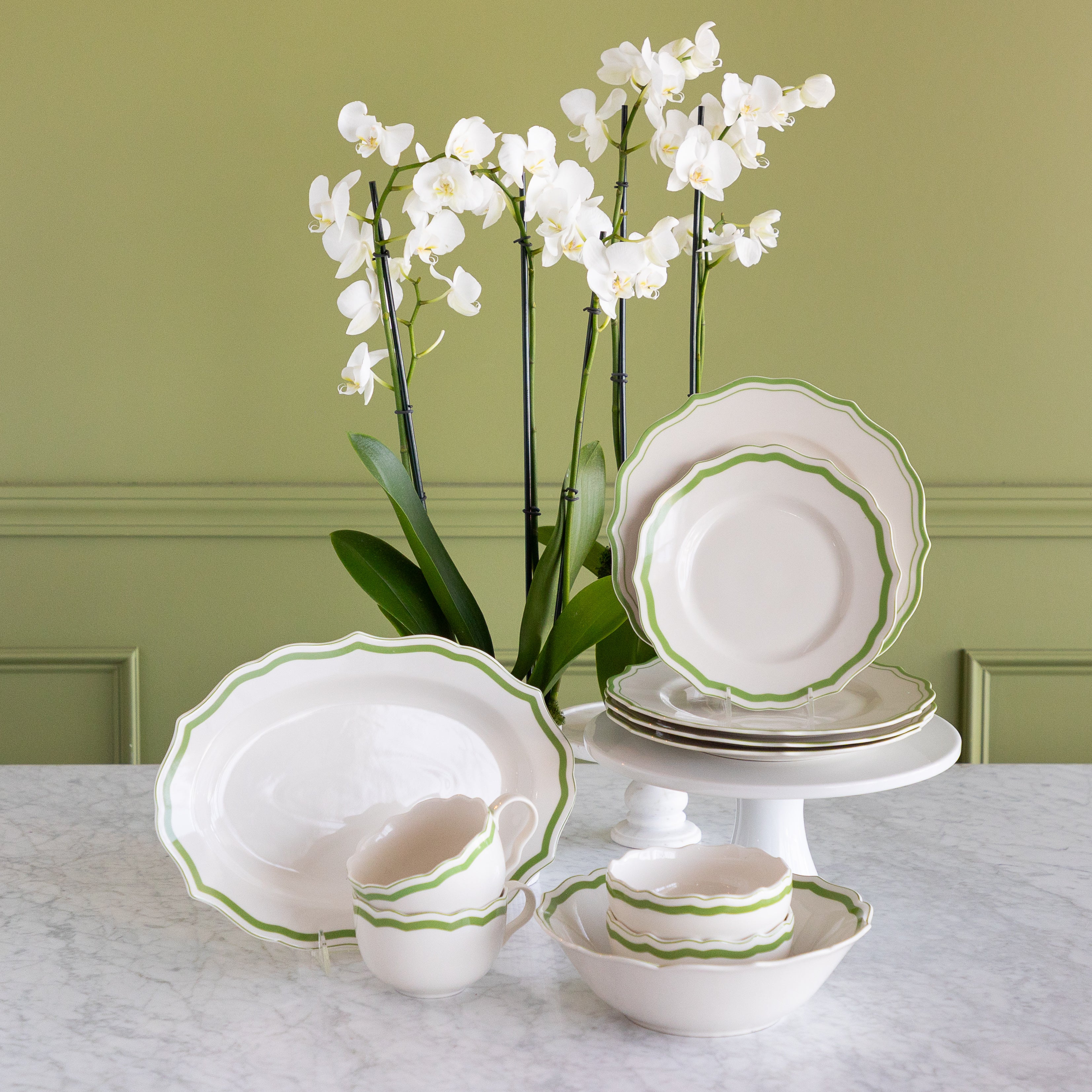 A coordinated white dinnerware set with a green scalloped rim, displayed on a marble table against an olive green paneled wall. The set includes plates, bowls, cups, and a platter, styled with white orchids in the background.