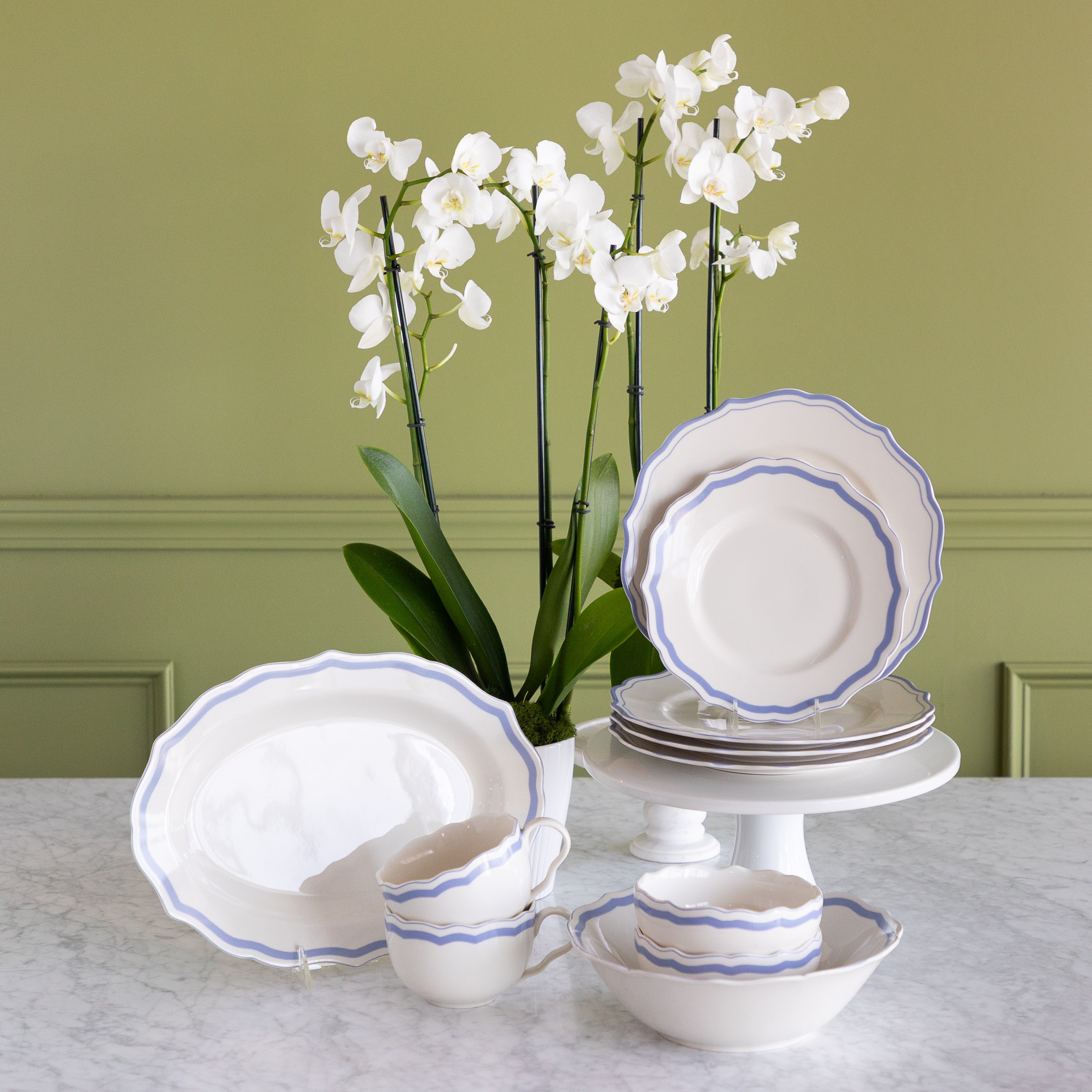 A white porcelain dinnerware set with blue scalloped rims, including plates, bowls, cups, and a platter, arranged on a marble table with white orchids against a sage green paneled wall.