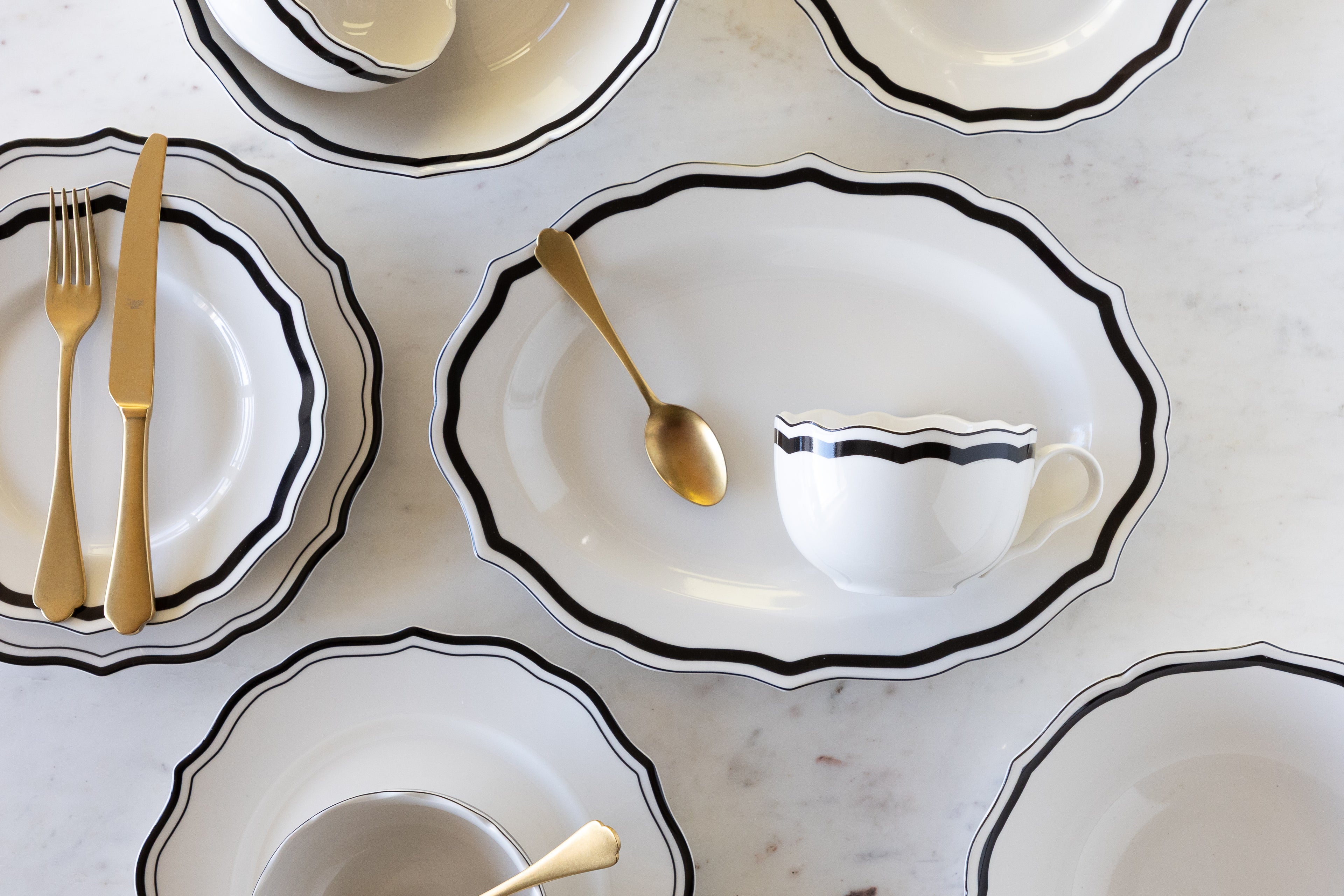 Set of white ceramic dishes with black rims and gold cutlery on a light gray surface.