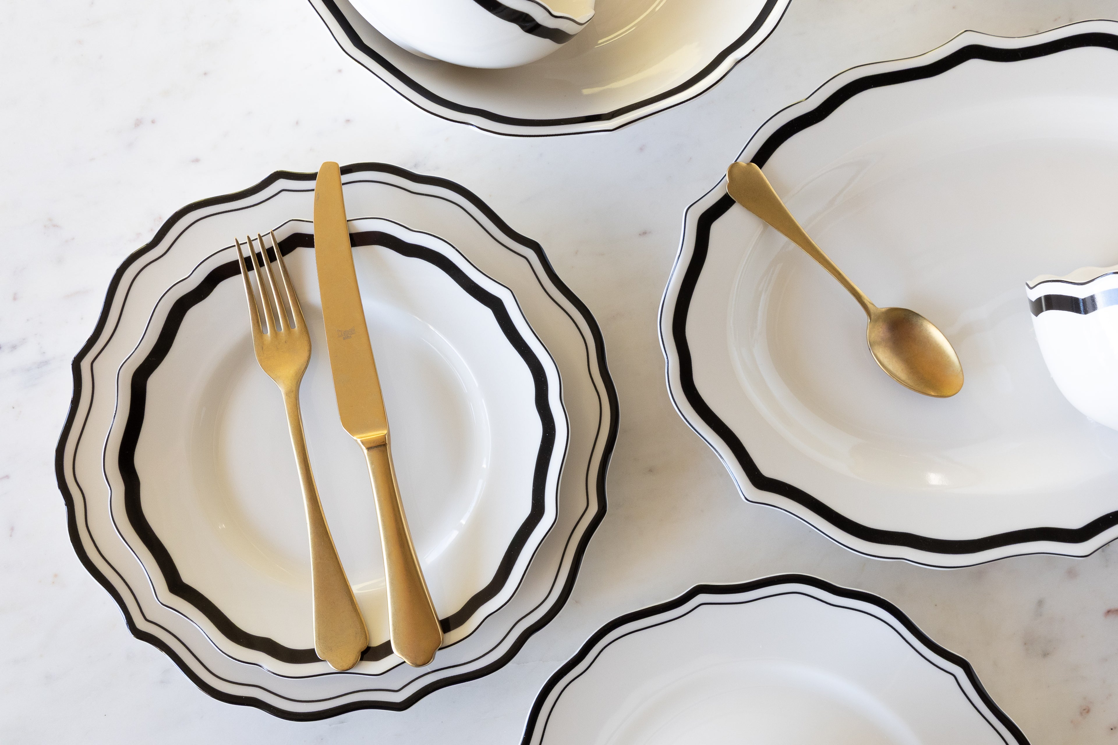Set of white ceramic dishes with black rims and gold cutlery on a light gray surface.