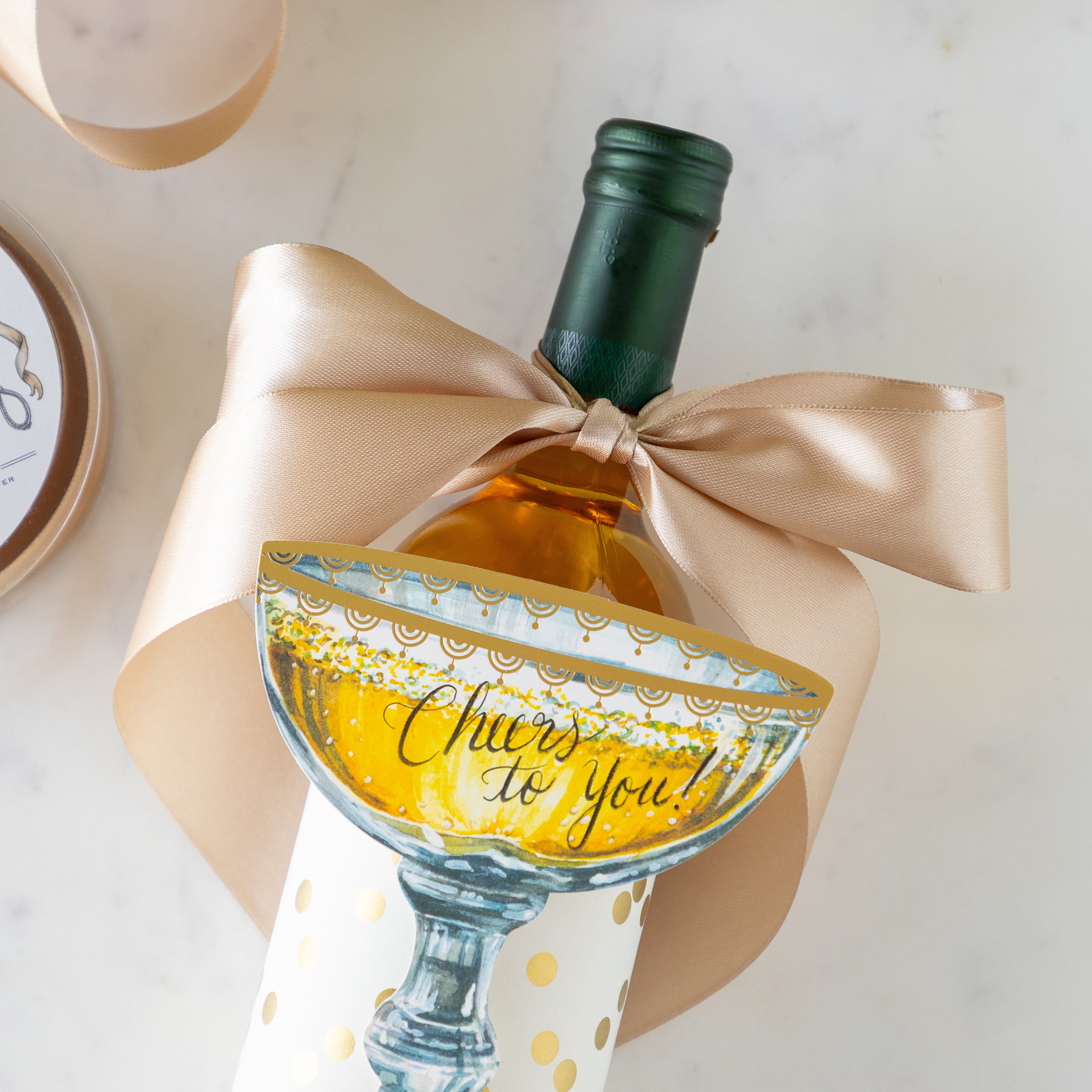 A bottle of wine with the Fancy Champagne Ribbon tied in a bow around it, wrapped in the Gold Confetti Placemat and Champagne Coupe Place Card atop reading "Cheers to You!".