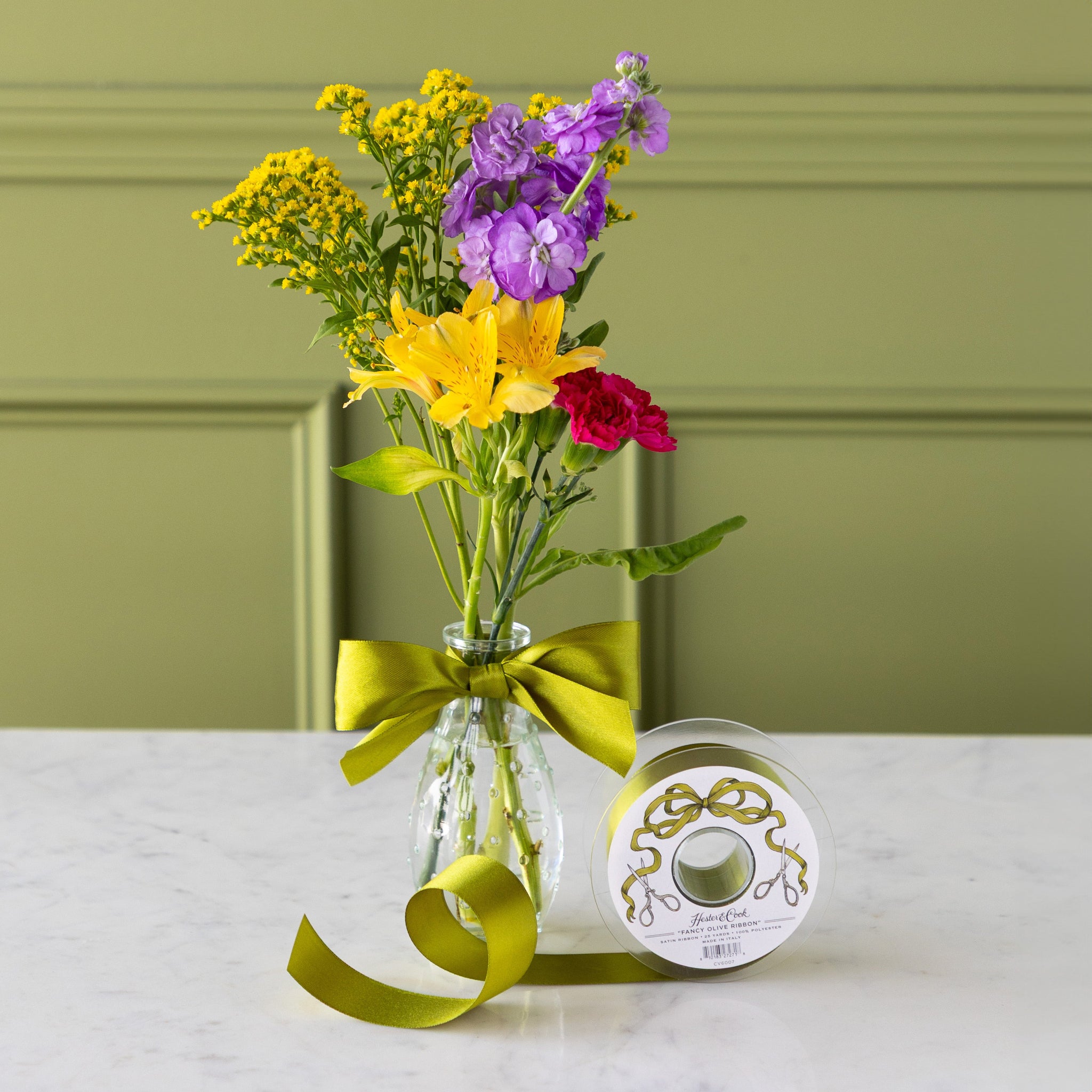 Vase of colorful flowers with a green ribbon bow tied around it, next to a spool of Fancy Olive Ribbon.