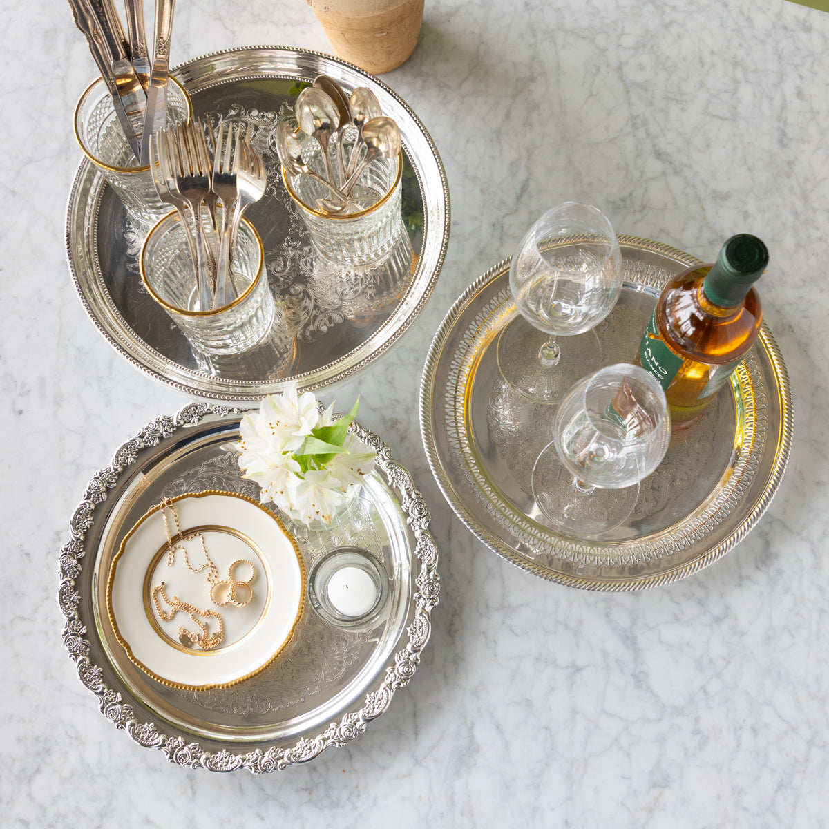 Decorative silver trays with jewelry, glasses, and a bottle on a marble surface.