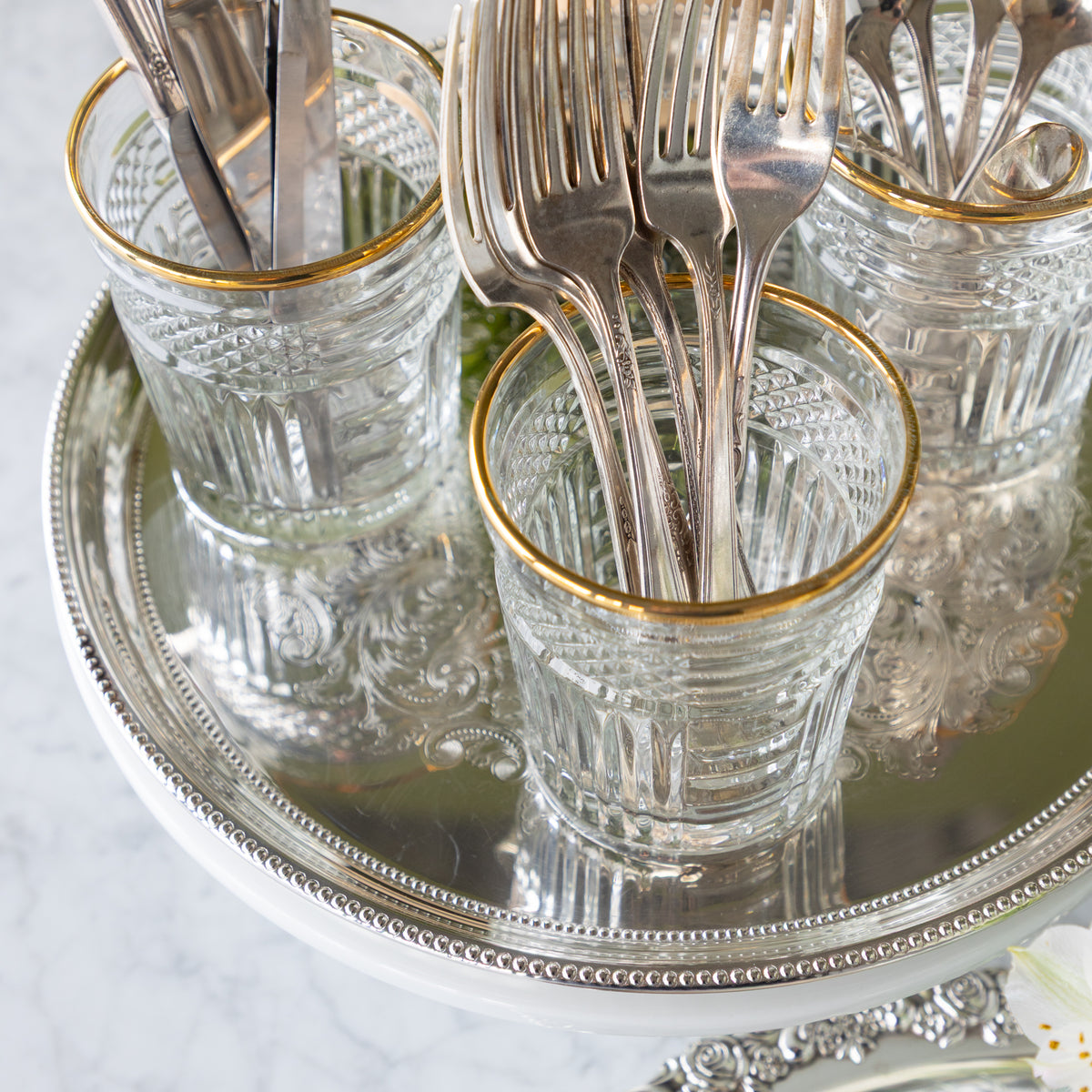 Clear glass cups with gold rims holding silver forks on a silver tray.