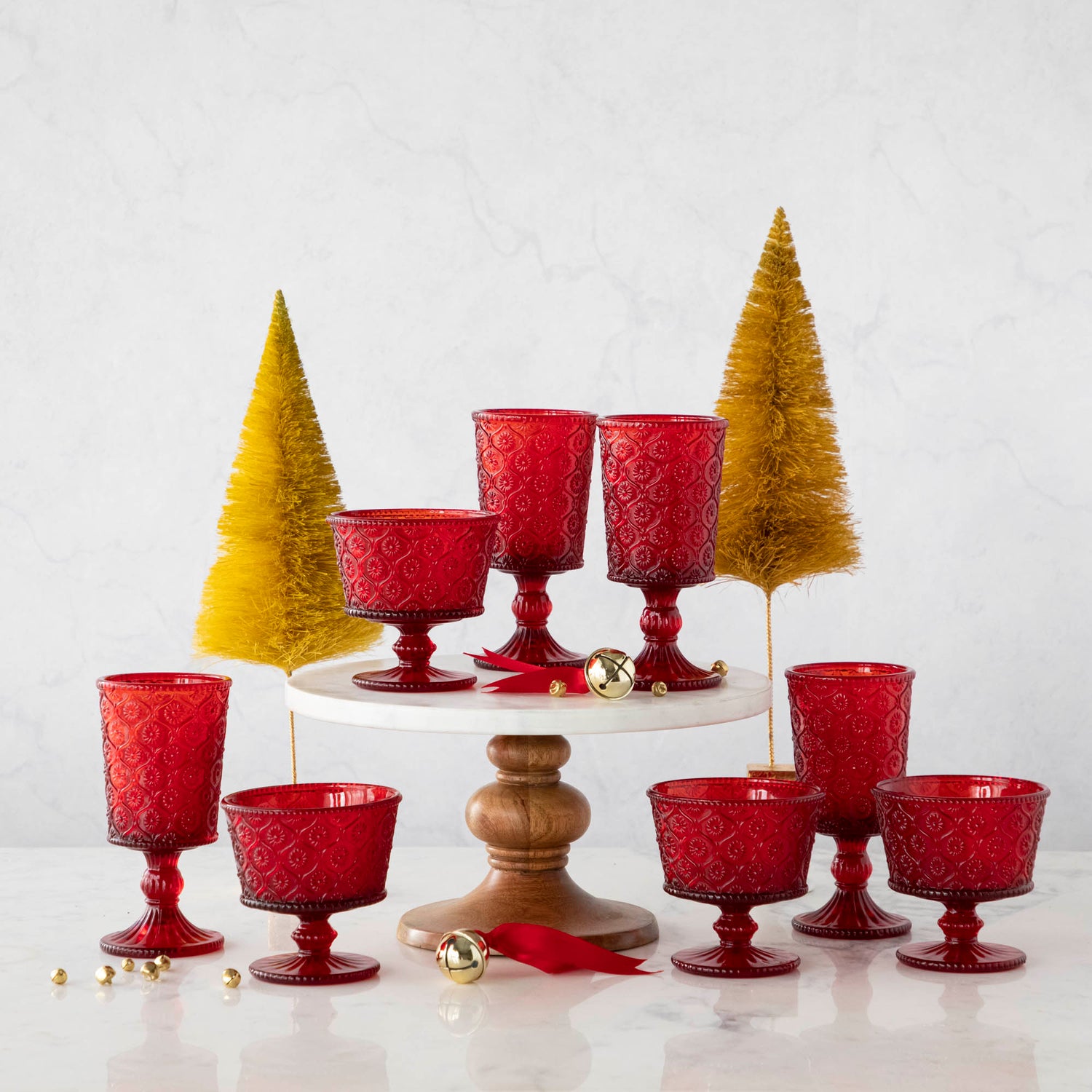 Elegant Qualia red pressed glassware set displayed on a white cake stand with wooden pedestal, flanked by green topiary balls on a marble background.