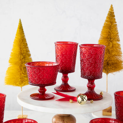 Elegant Qualia red pressed glassware set displayed on a white cake stand with wooden pedestal, flanked by green topiary balls on a marble background.