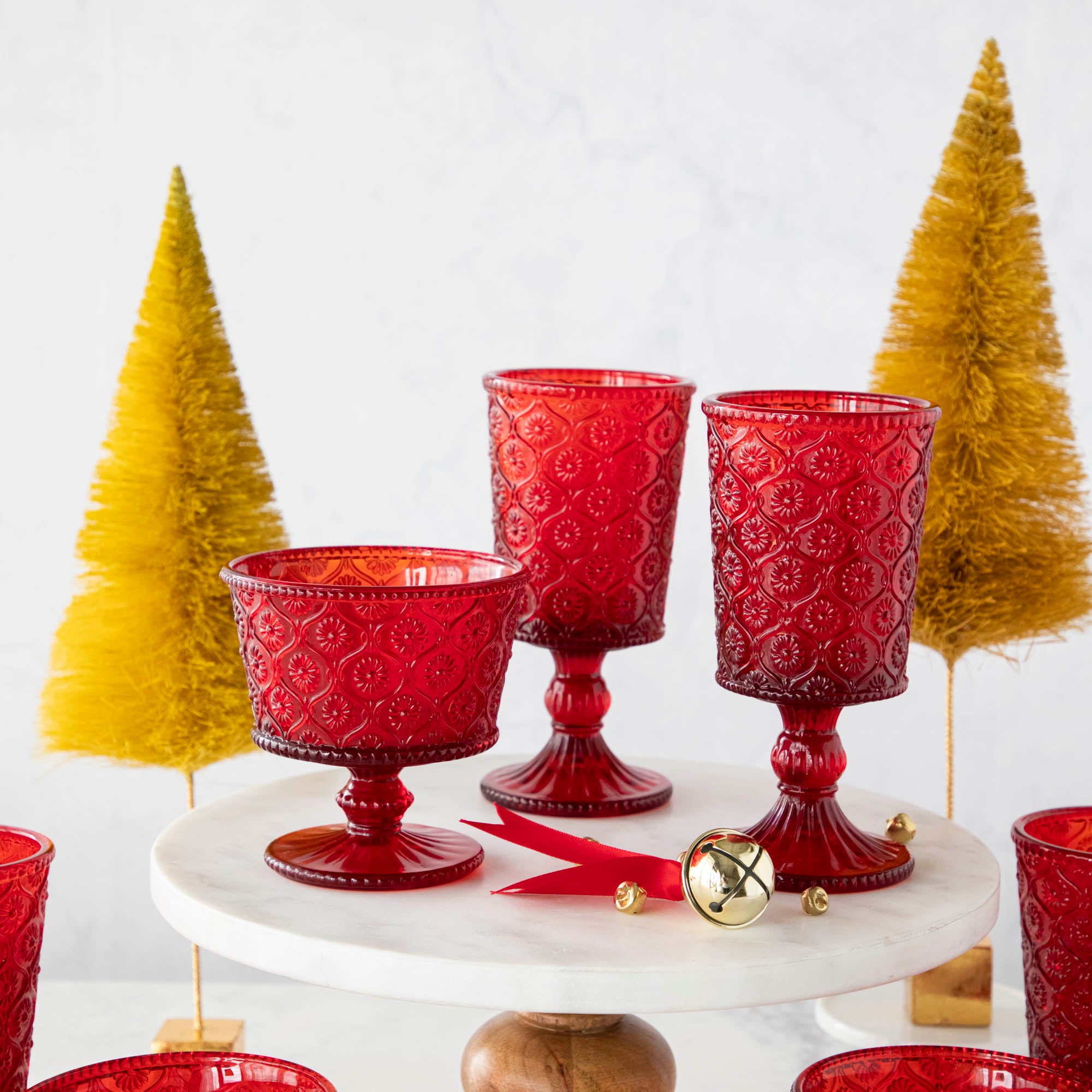 Elegant Qualia red pressed glassware set displayed on a white cake stand with wooden pedestal, surrounded by gold flocked trees and scattered jingle bells.