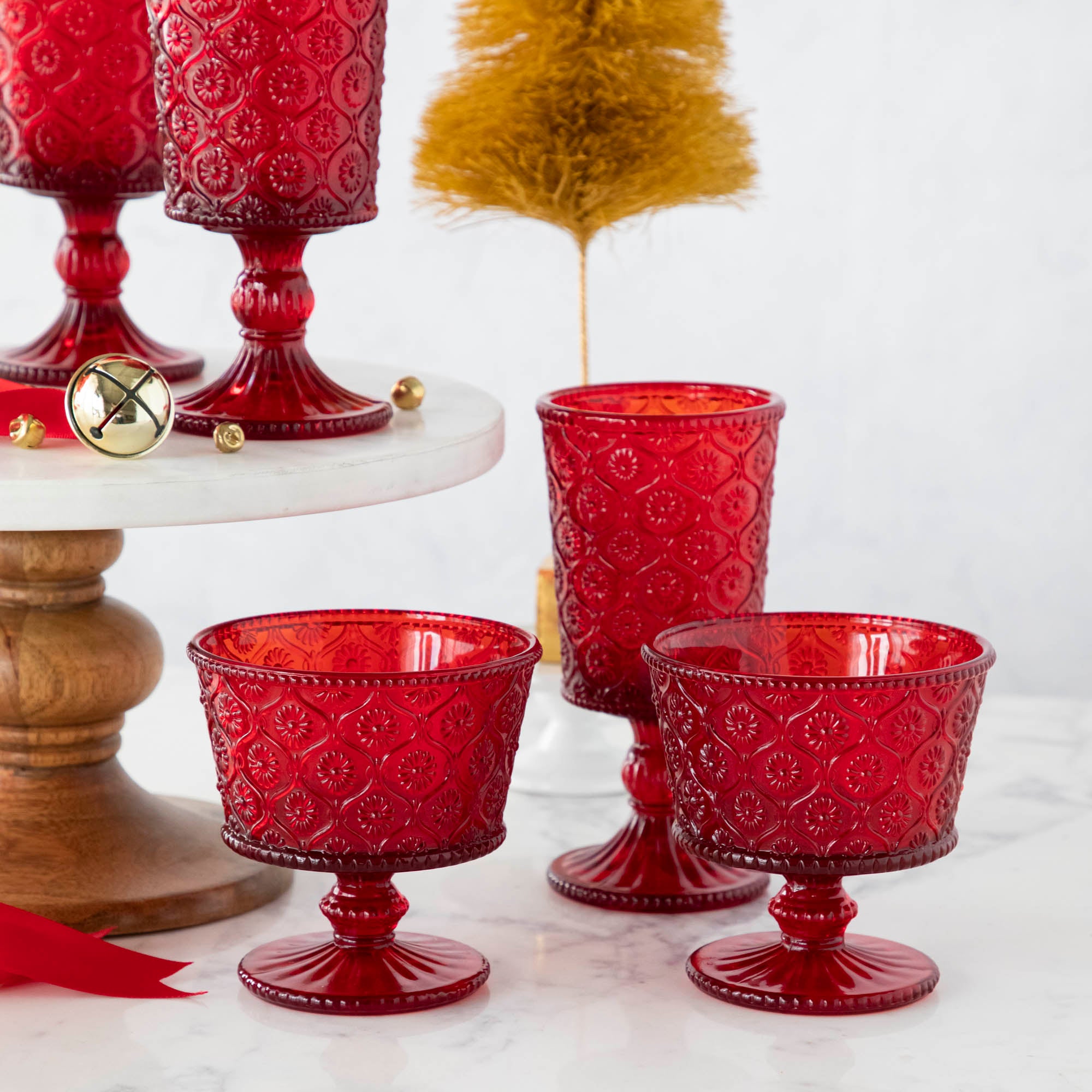 Elegant Qualia red pressed glassware set displayed on a white cake stand with wooden pedestal, surrounded by gold flocked trees and scattered jingle bells.