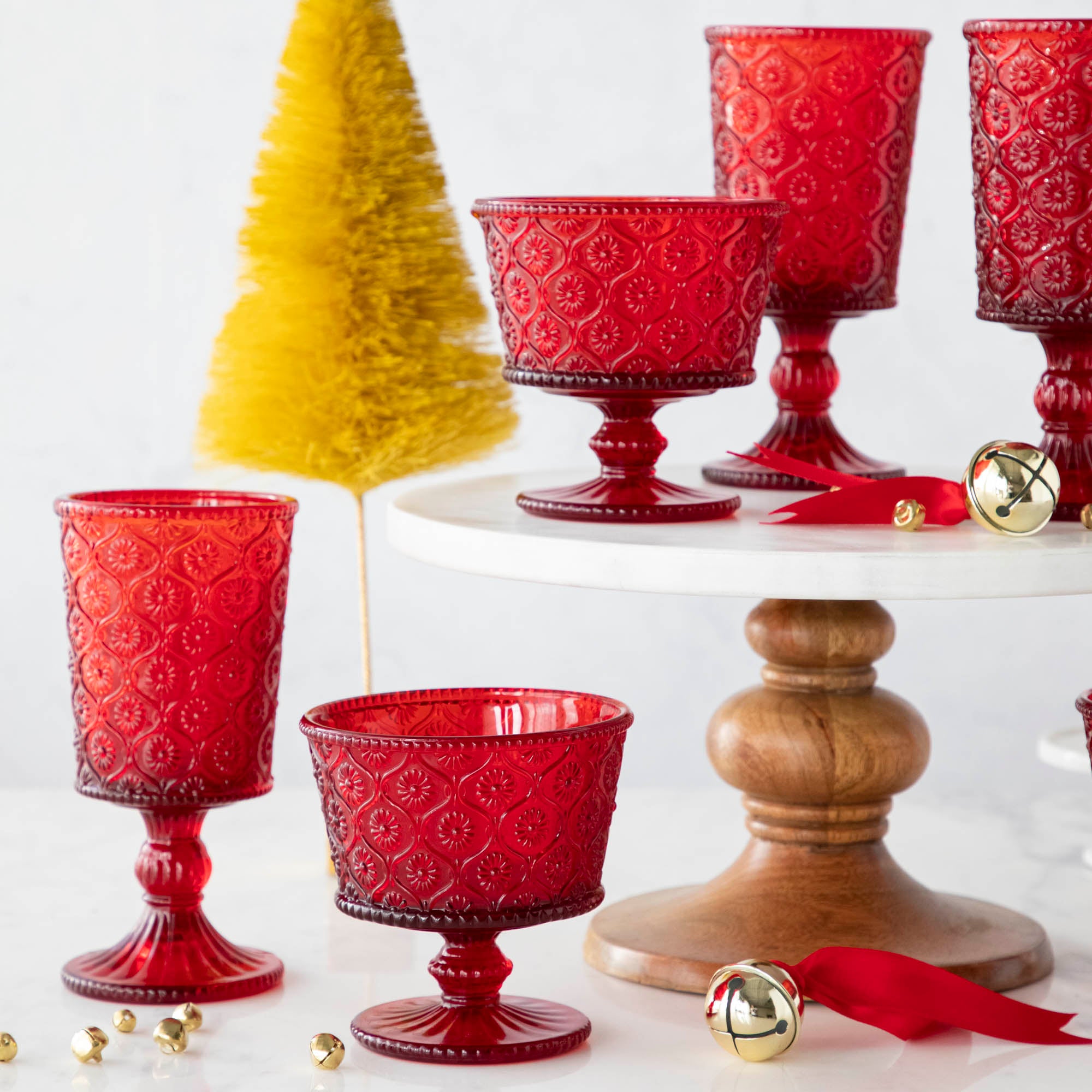 Elegant Qualia red pressed glassware set displayed on a white cake stand with wooden pedestal, surrounded by gold flocked trees and scattered jingle bells.