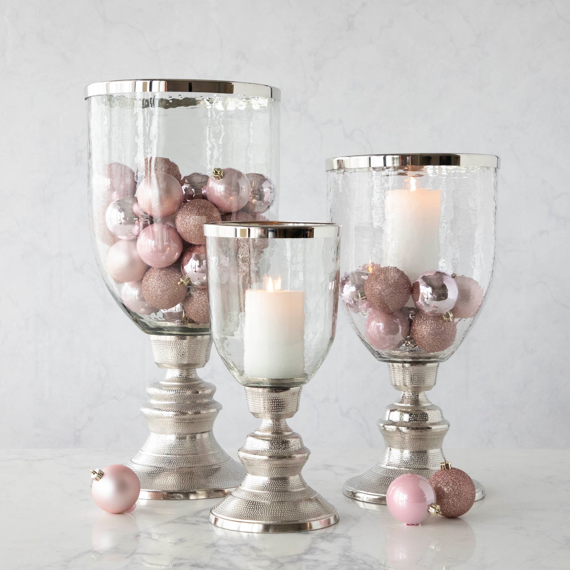 A small, medium and large Candle Hurricane Holder with a candle and pink ornaments.