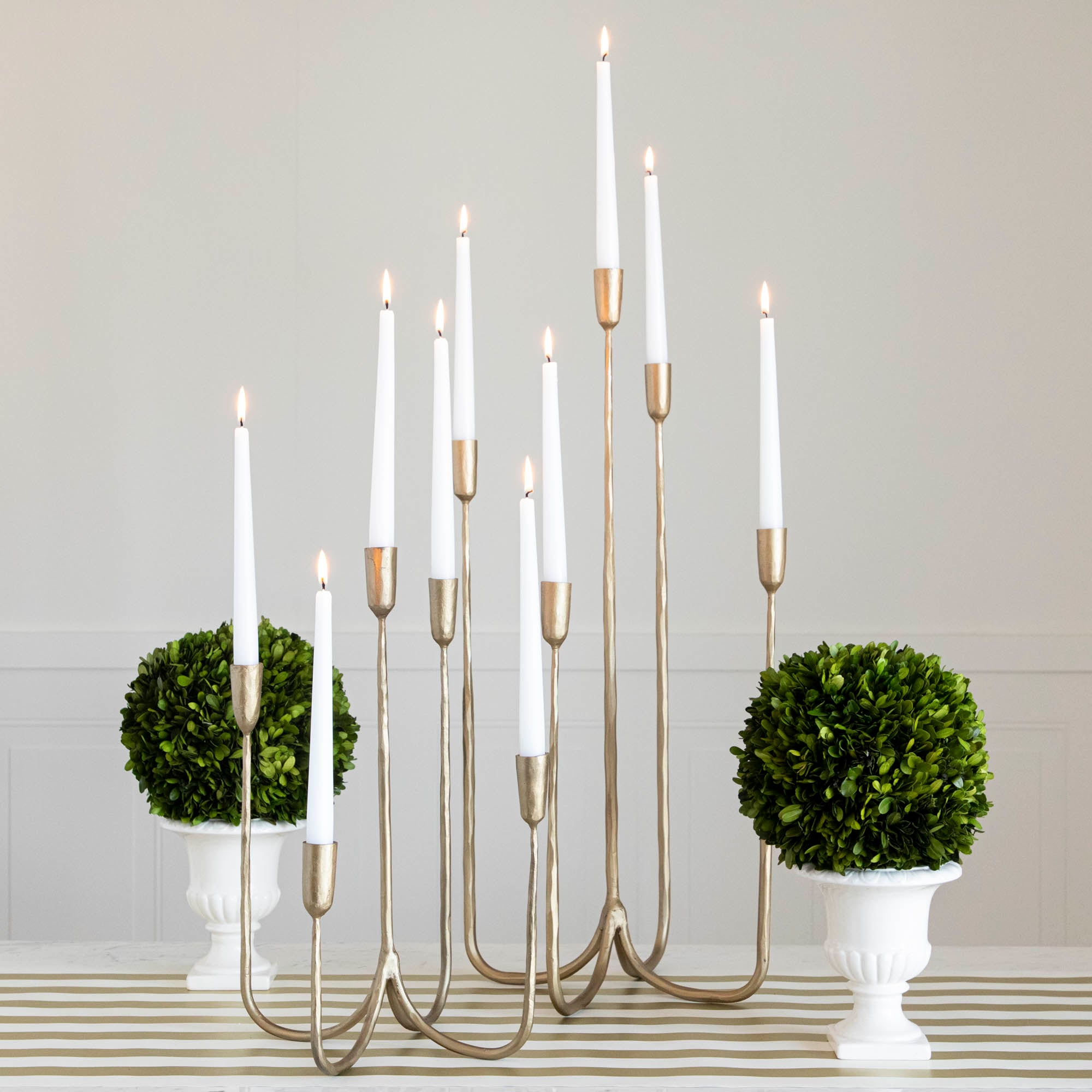 Forged Brass Candelabra in short and tall sizes with white candles lit in them on a table with vases that have boxwood balls on them.