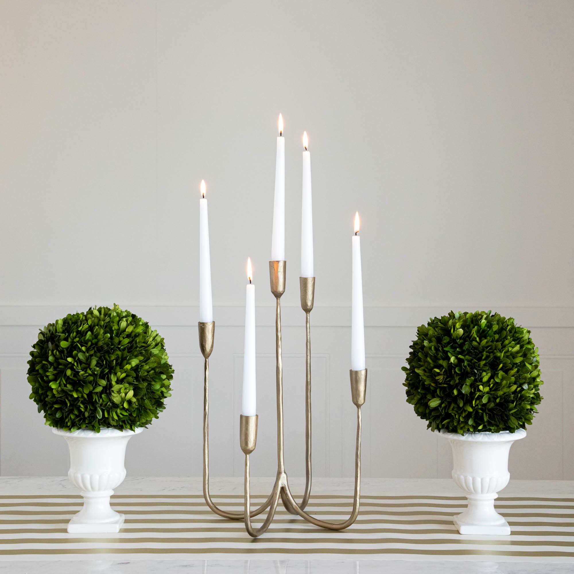 Forged Brass Candelabra in short size with white candles lit in it on a table with vases that have boxwood balls on them.