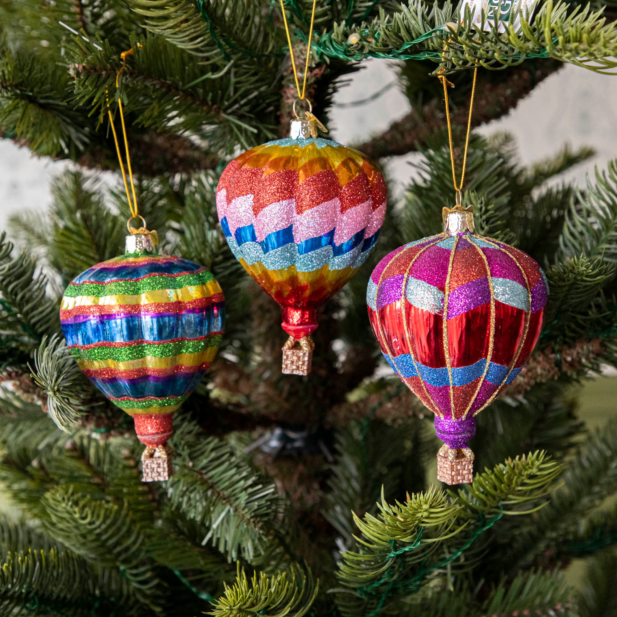 A set of three glass hot air balloon ornaments in red, blue, and green stripes, hanging on a Christmas tree.