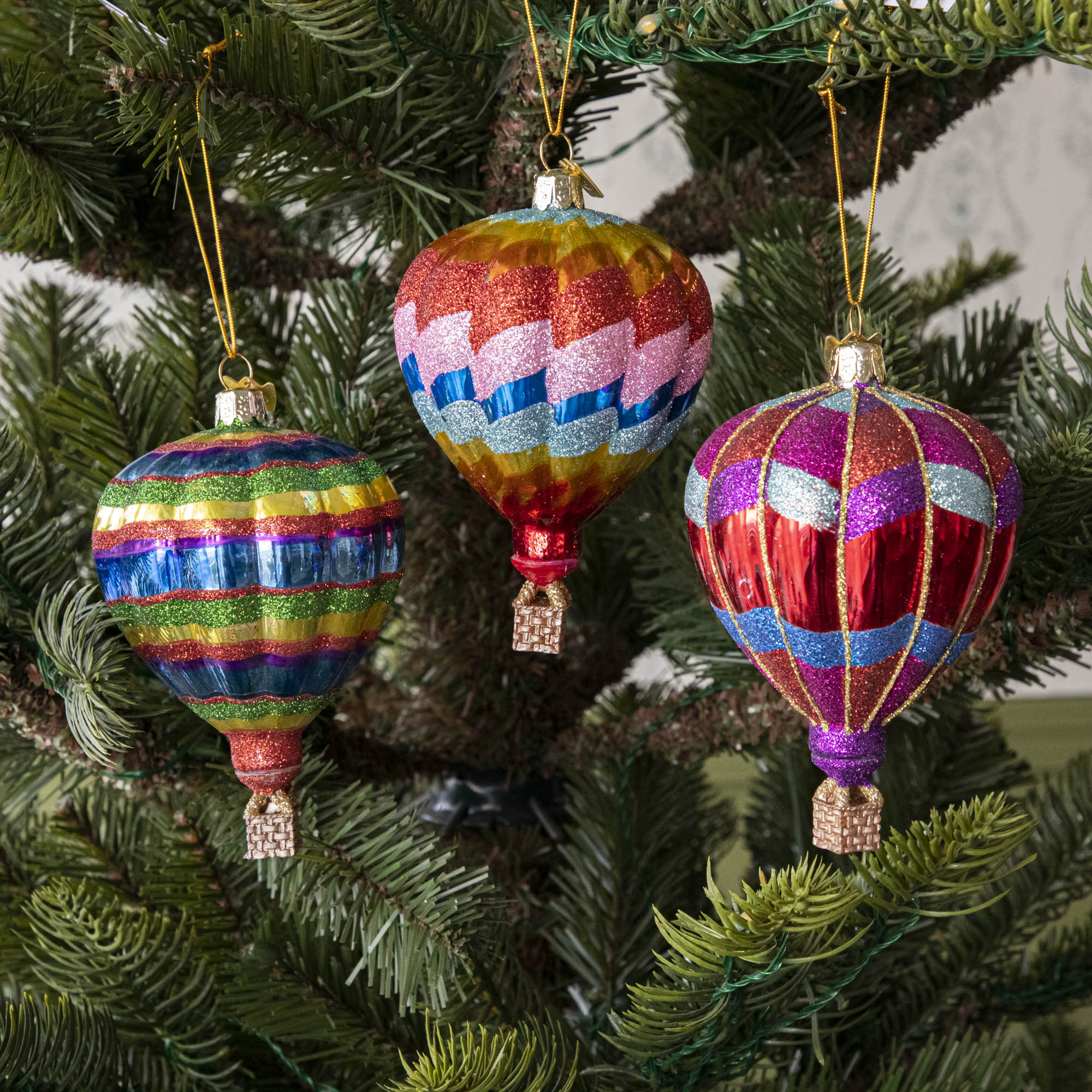 A set of three glass hot air balloon ornaments in red, blue, and green stripes, hanging on a Christmas tree.
