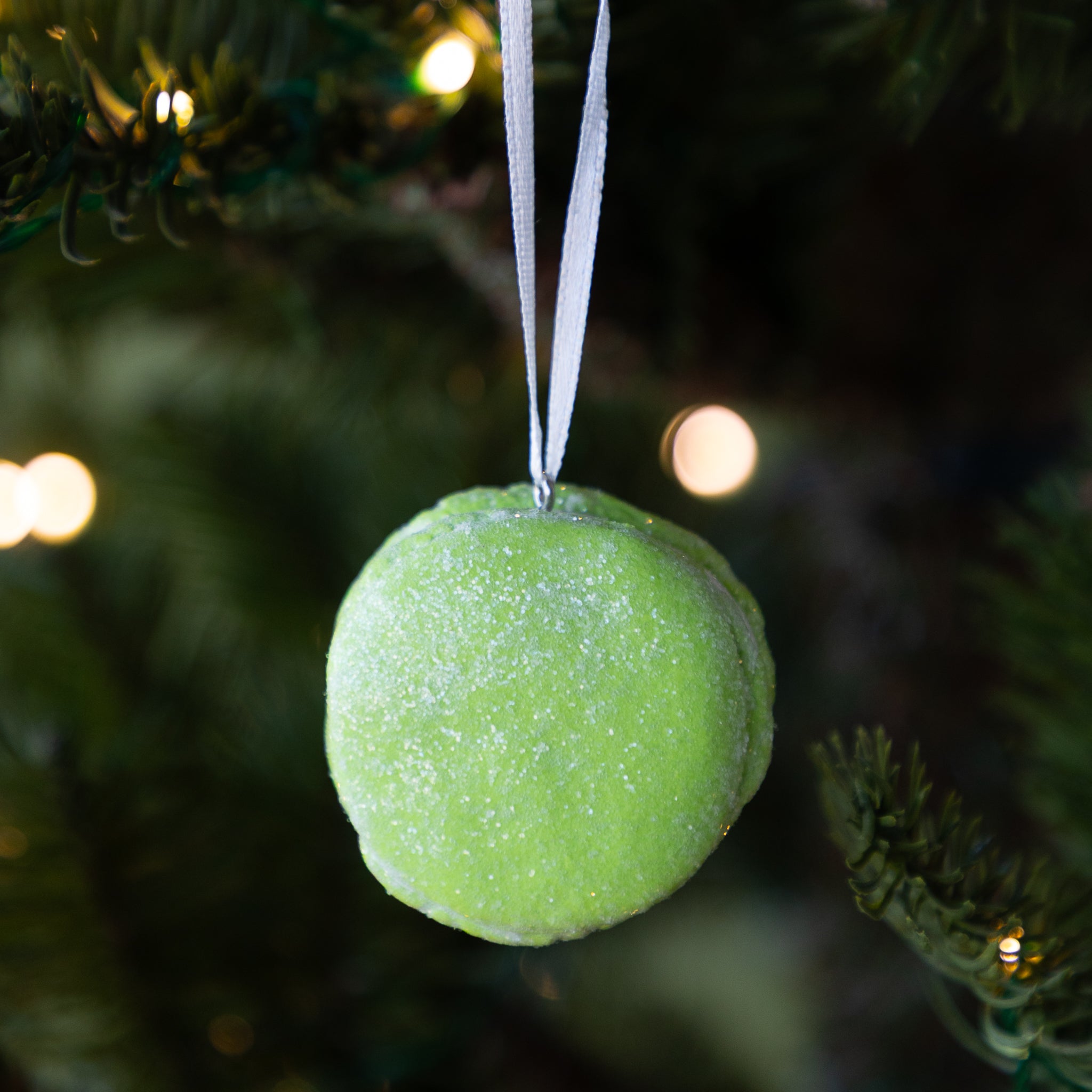 Green macaron ornament hanging on a Christmas tree with blurred lights in the background.