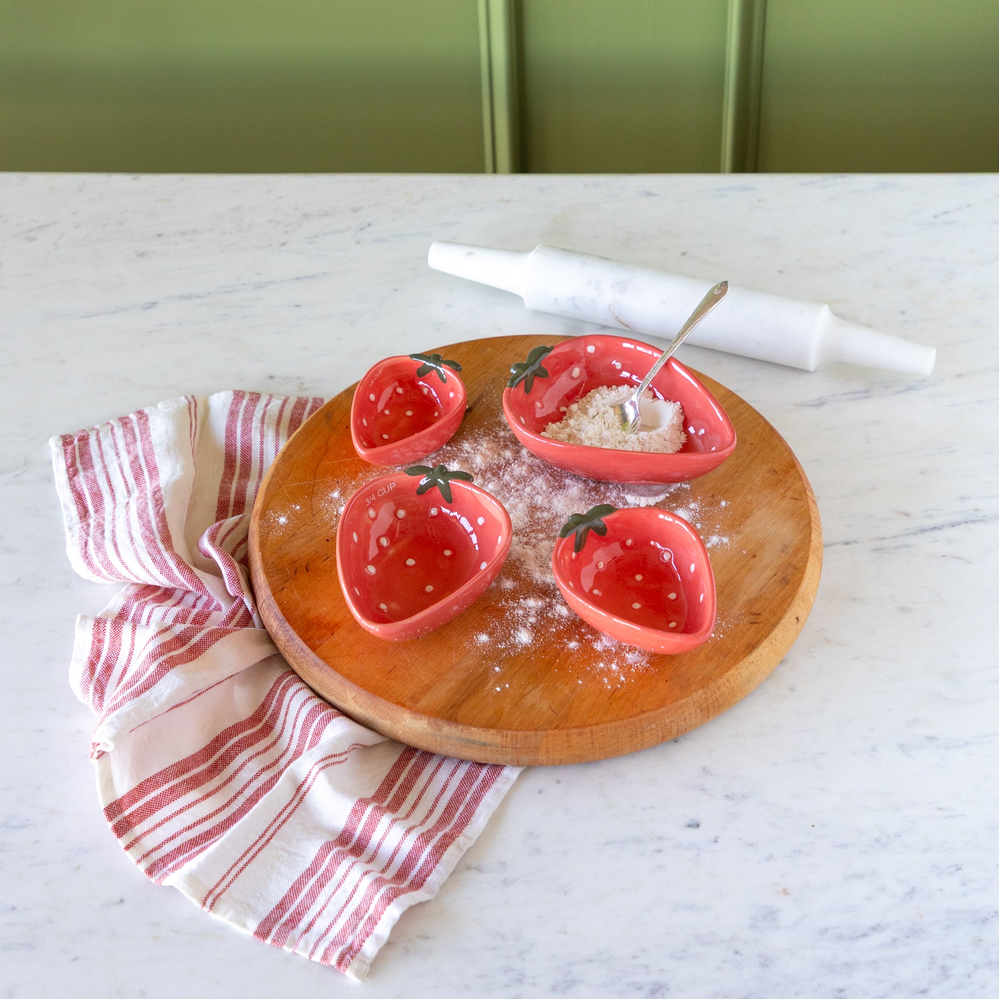 Wooden board with Strawberry Shaped Measuring Cups, a red-and-white linen napkin and a marble roller, on a marble surface.