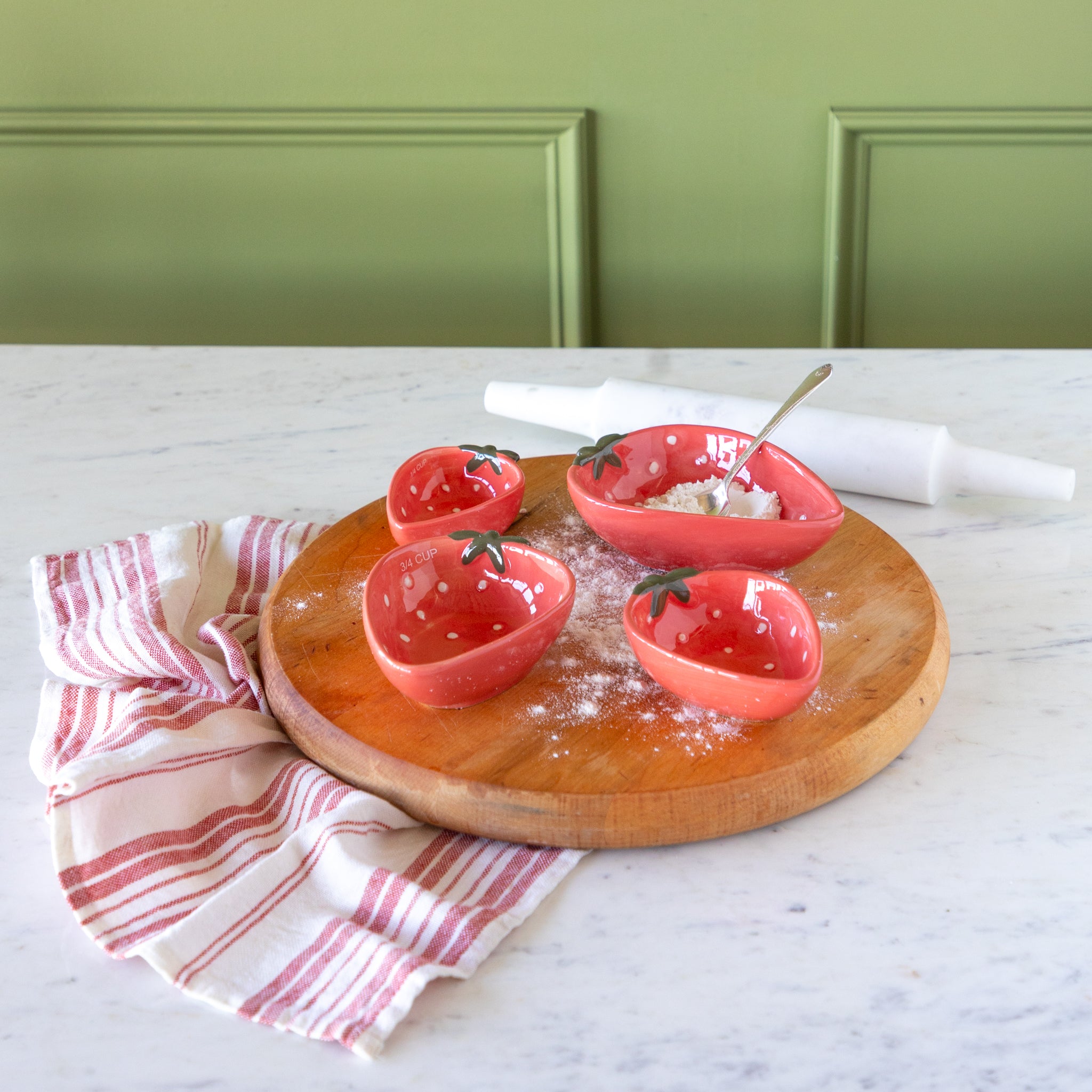 Wooden board with Strawberry Shaped Measuring Cups, a red-and-white linen napkin and a marble roller, on a marble surface.