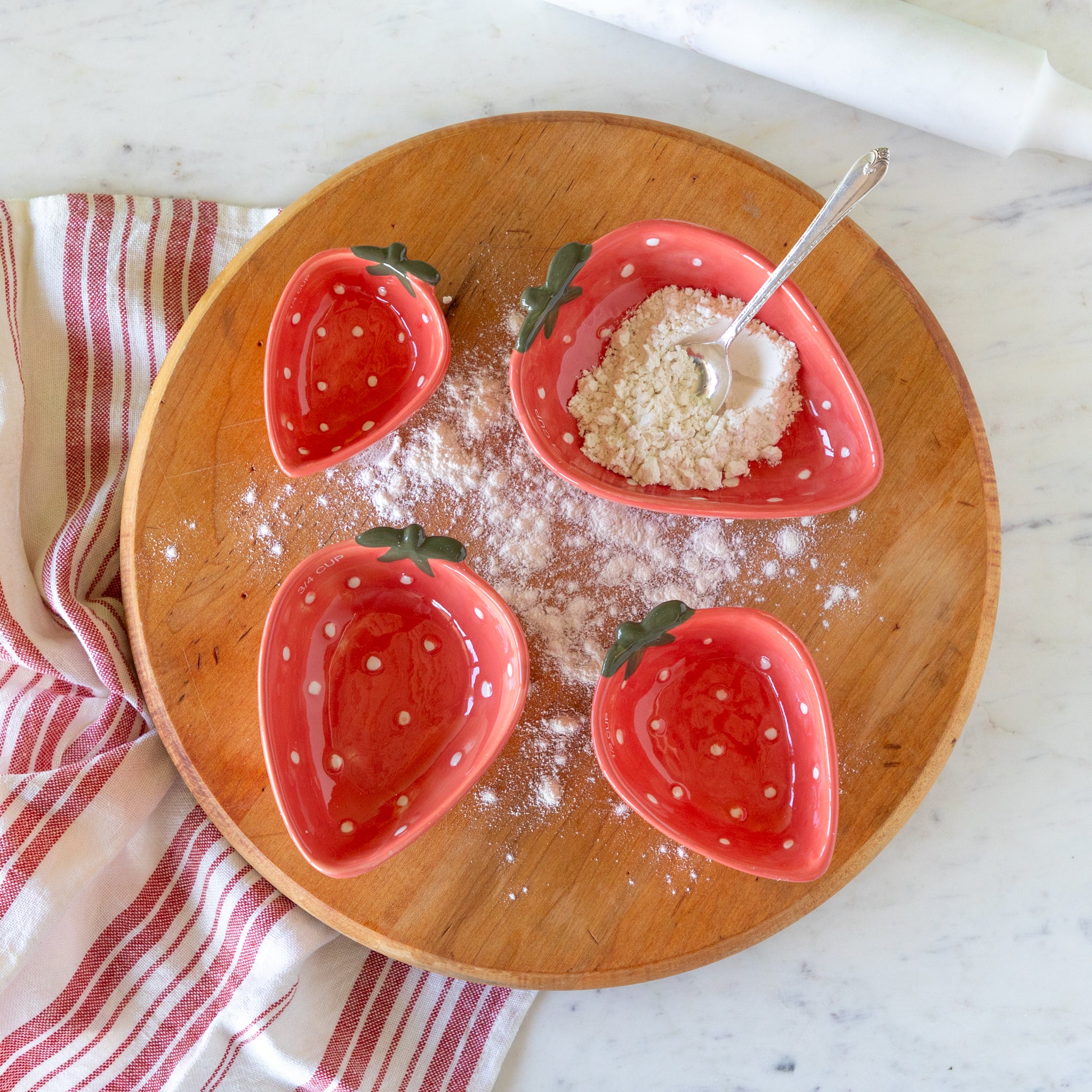 Red strawberry-shaped measuring cups on a wooden surface with a spoon and flour.