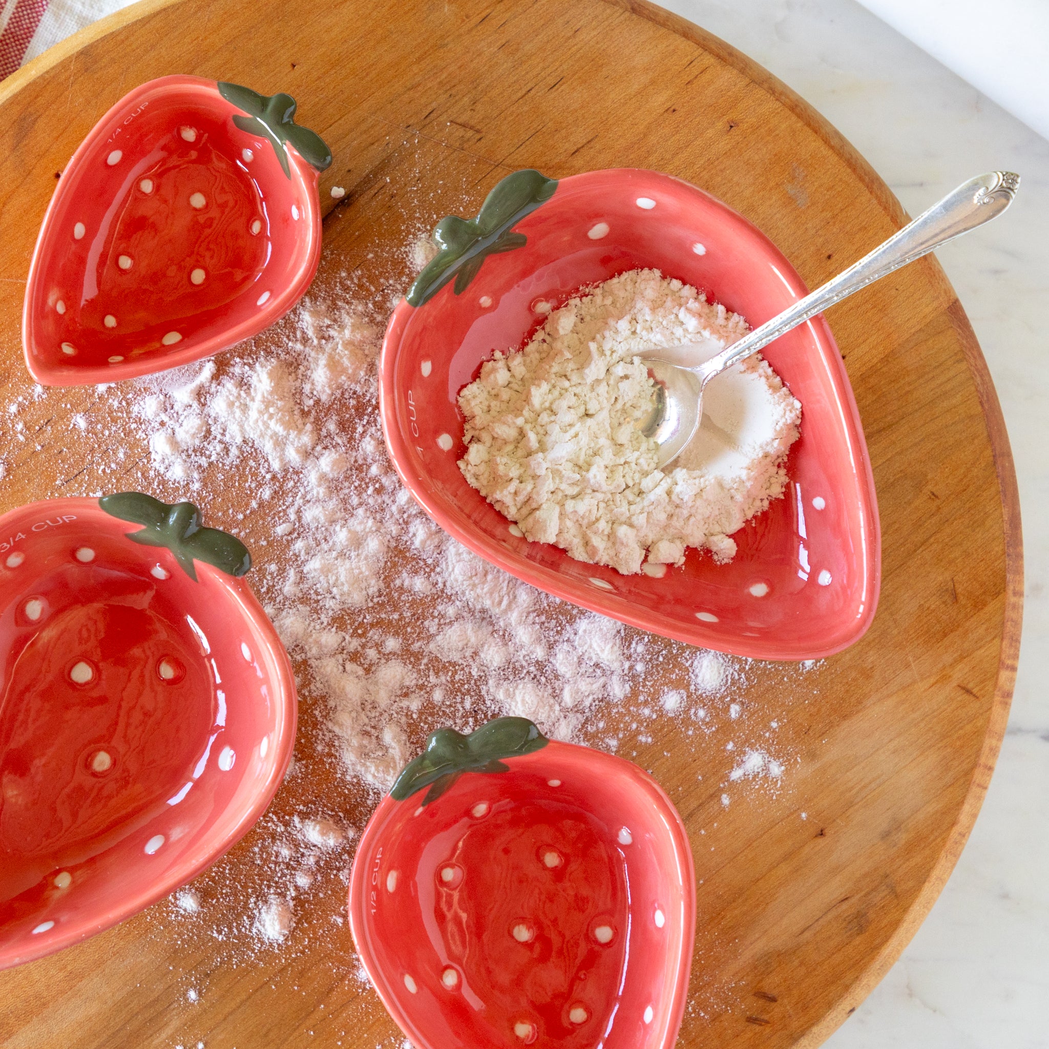 Red strawberry-shaped measuring cups on a wooden surface with a spoon and flour.