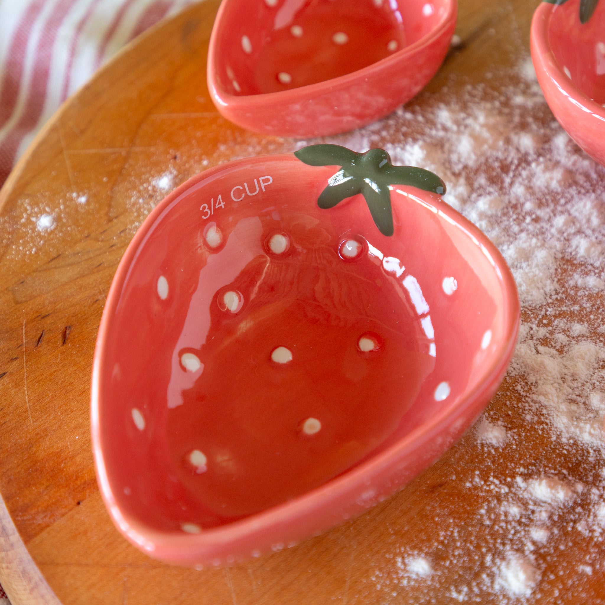 Red ceramic bowl measuring cup shaped like a strawberry on a wooden surface with flour.