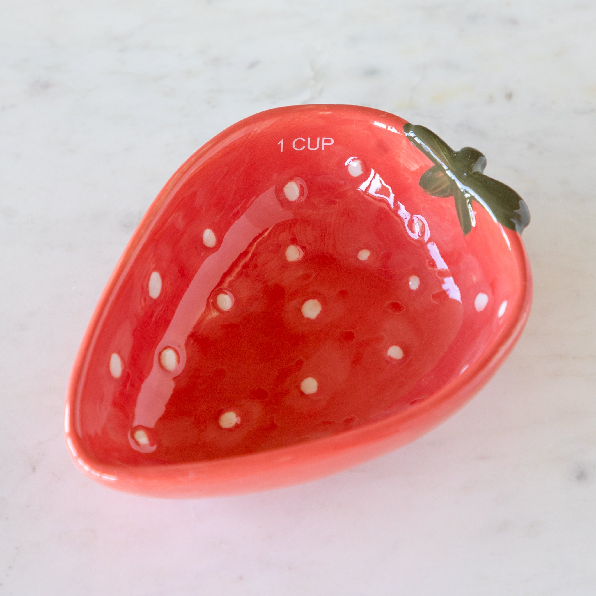 Red ceramic measuring cup shaped like a strawberry with white speckles, on a white surface.