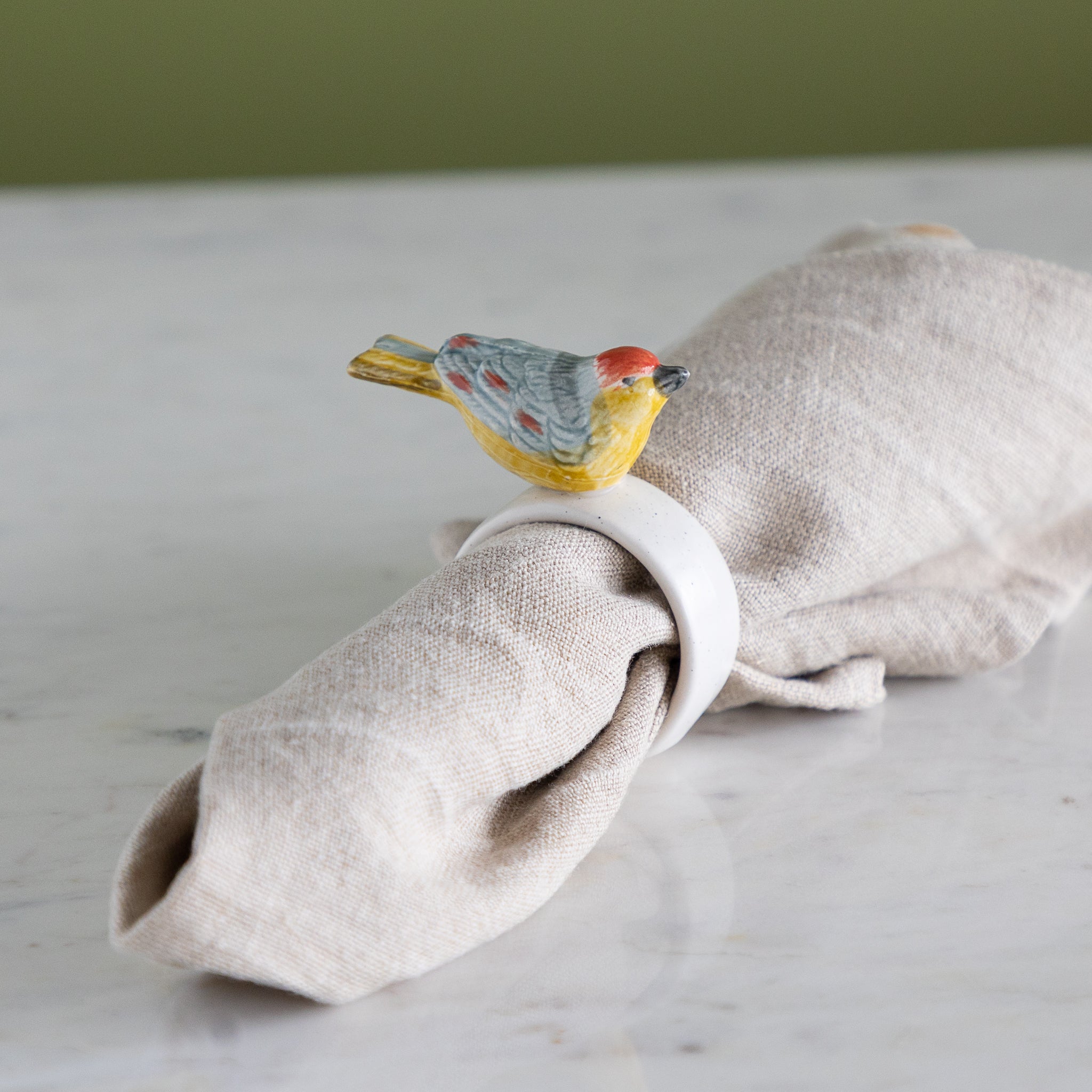 Napkin ring with colorful bird design around a folded beige napkin against a neutral background.