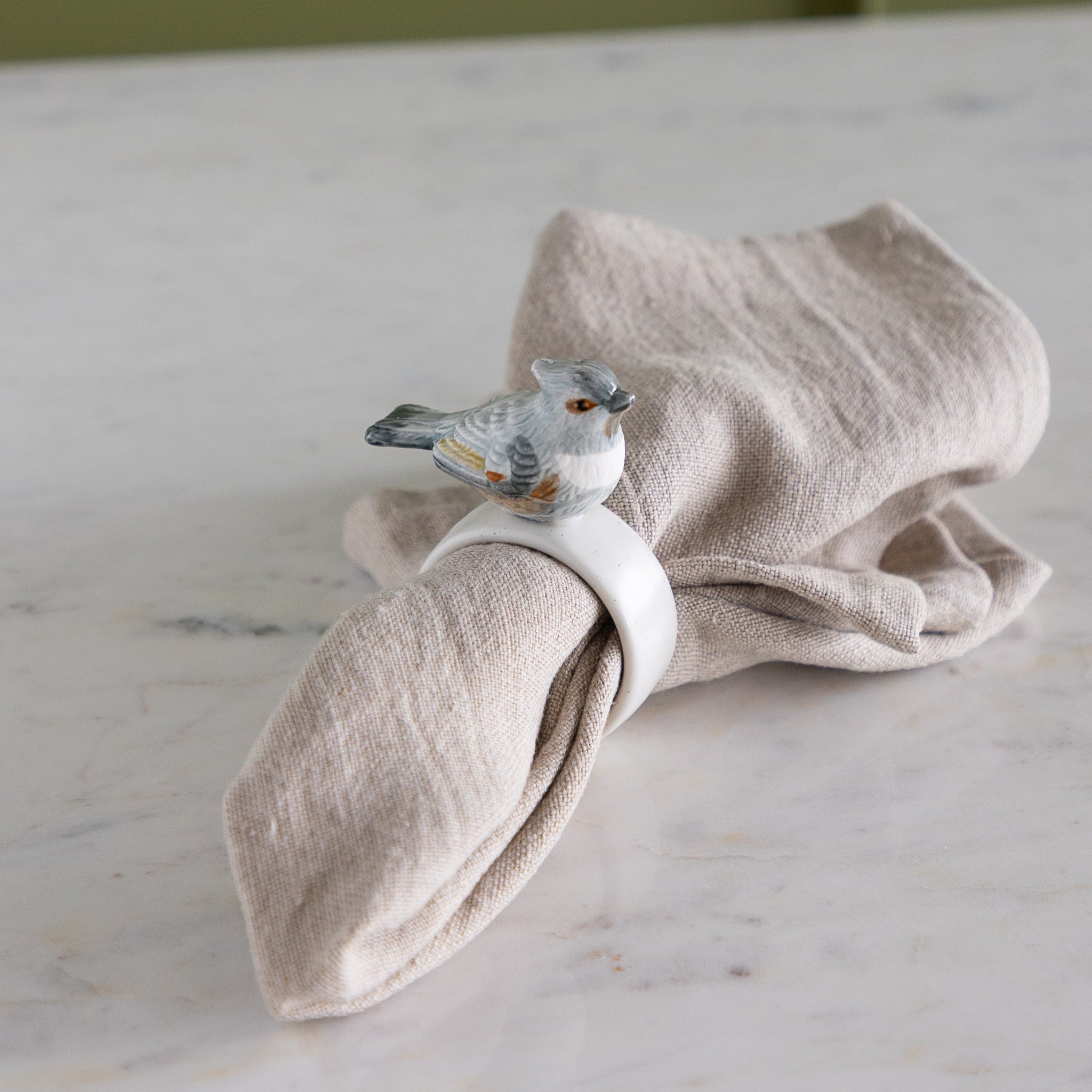 Napkin ring with grayish-blue bird design around a folded beige napkin against a neutral background.
