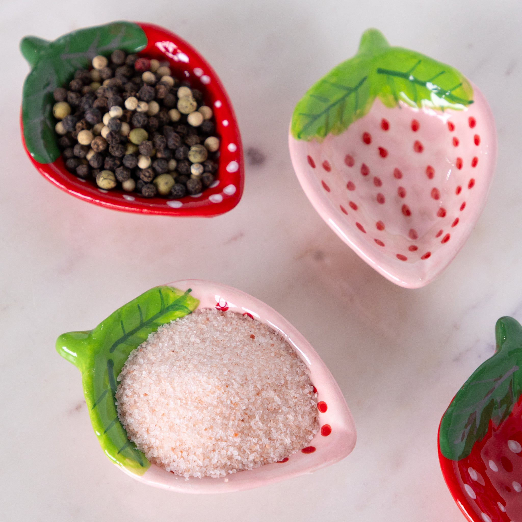 Strawberry-shaped ceramic containers with black and white peppercorns and sugar on a marble surface.