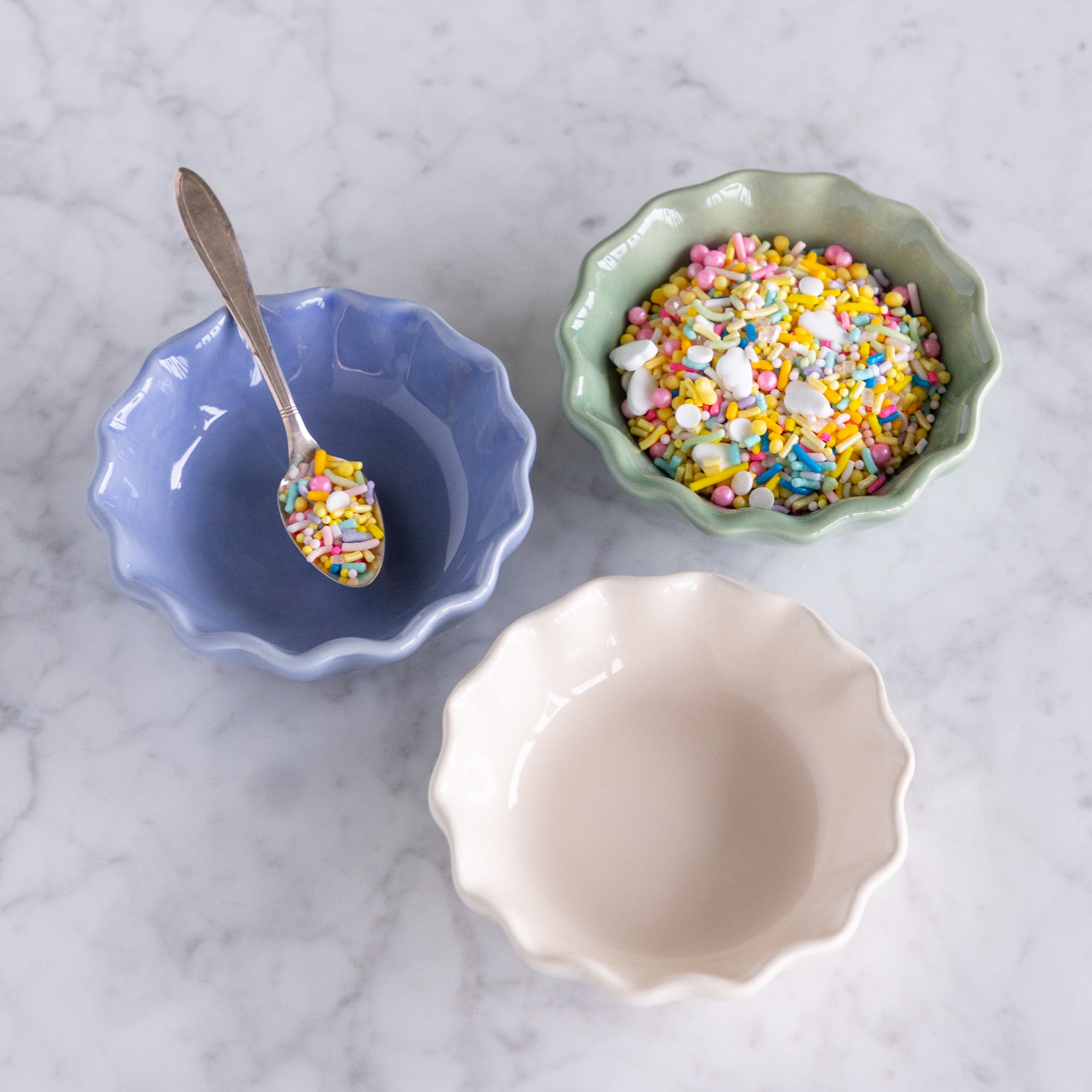 Three ceramic bowls in green, white and cream with colorful sprinkles on a marble surface.