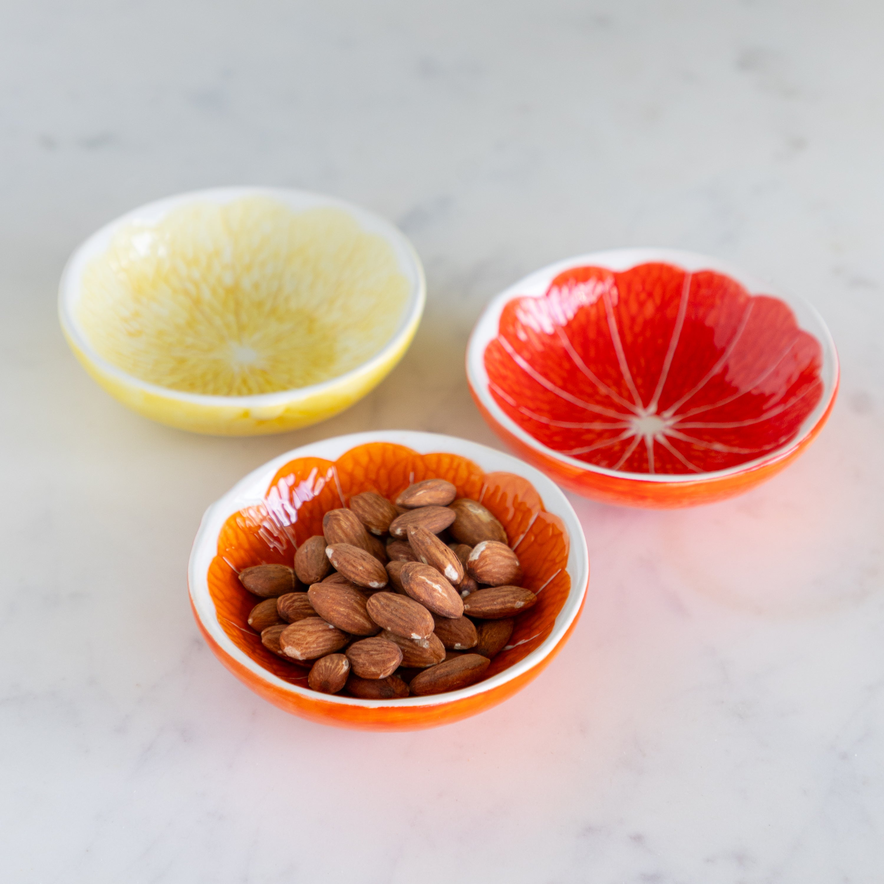Three Ceramic Citrus Fruit Shaped Bowls ––one filled with almonds on a marble table.