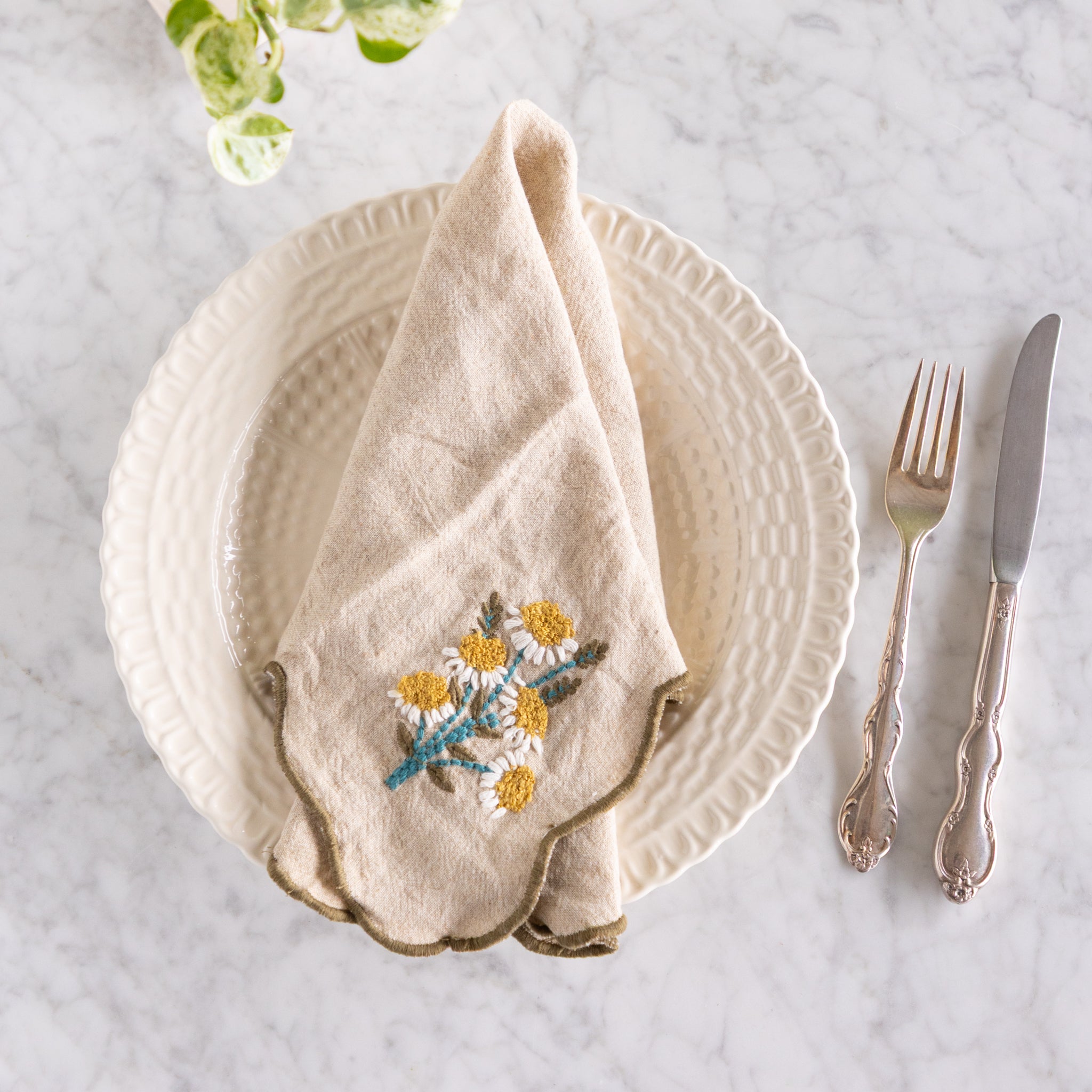 Beige embroidered daisies napkin on a white plate with silverware on a marble surface.