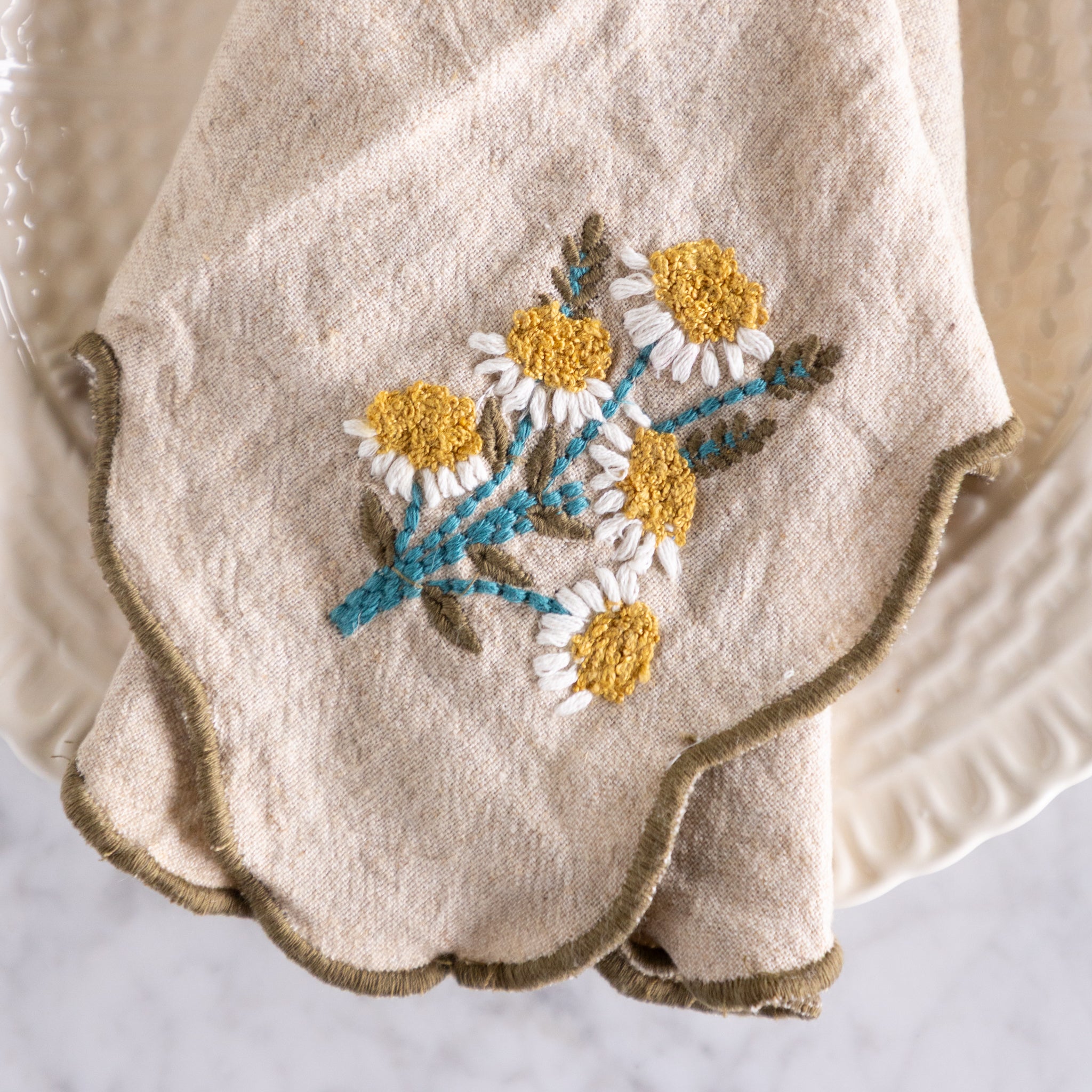 Embroidered daisies design on a beige napkin with a scalloped edge.