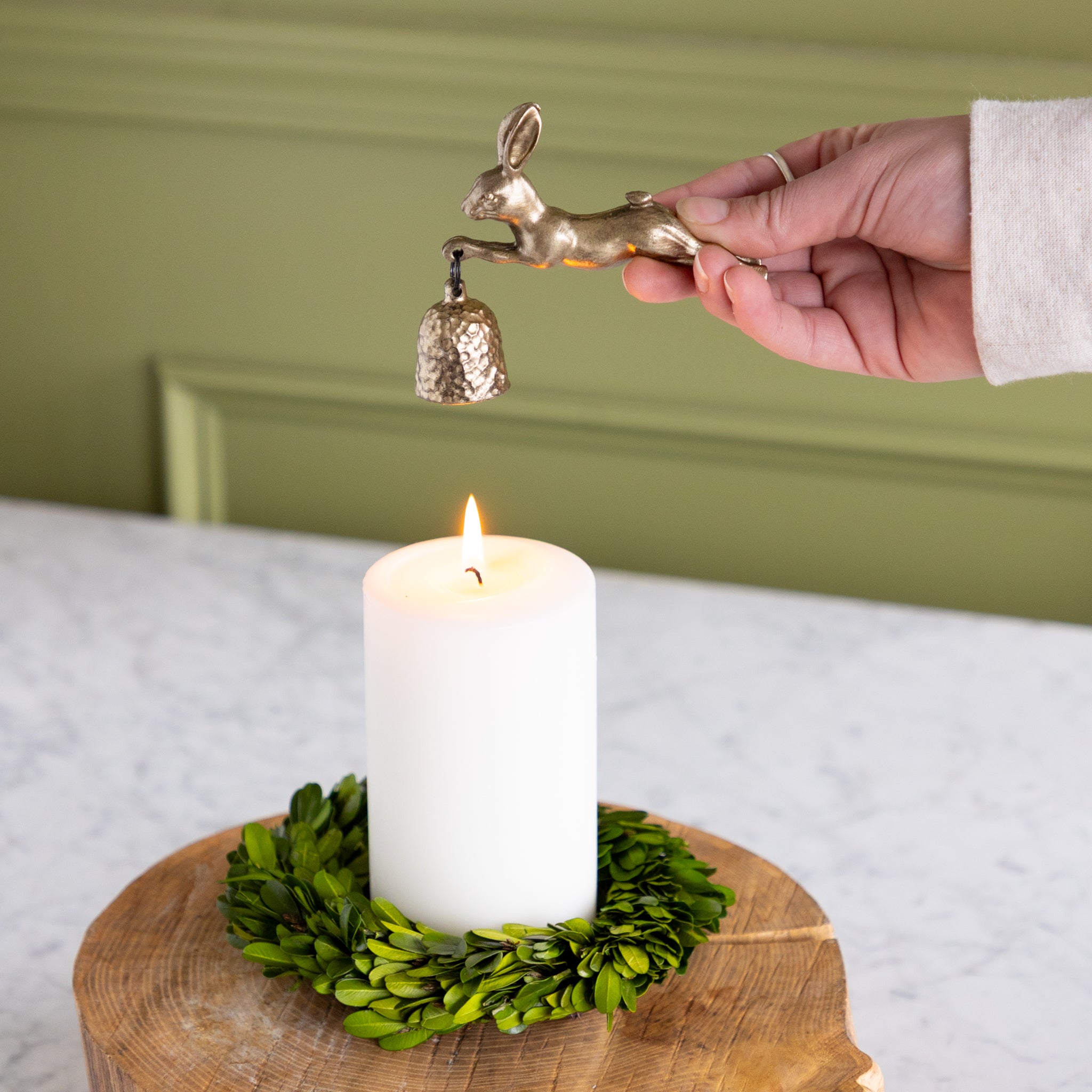 Hand holding a gold hare-shaped candle snuffer over a white candle on a green wreath.