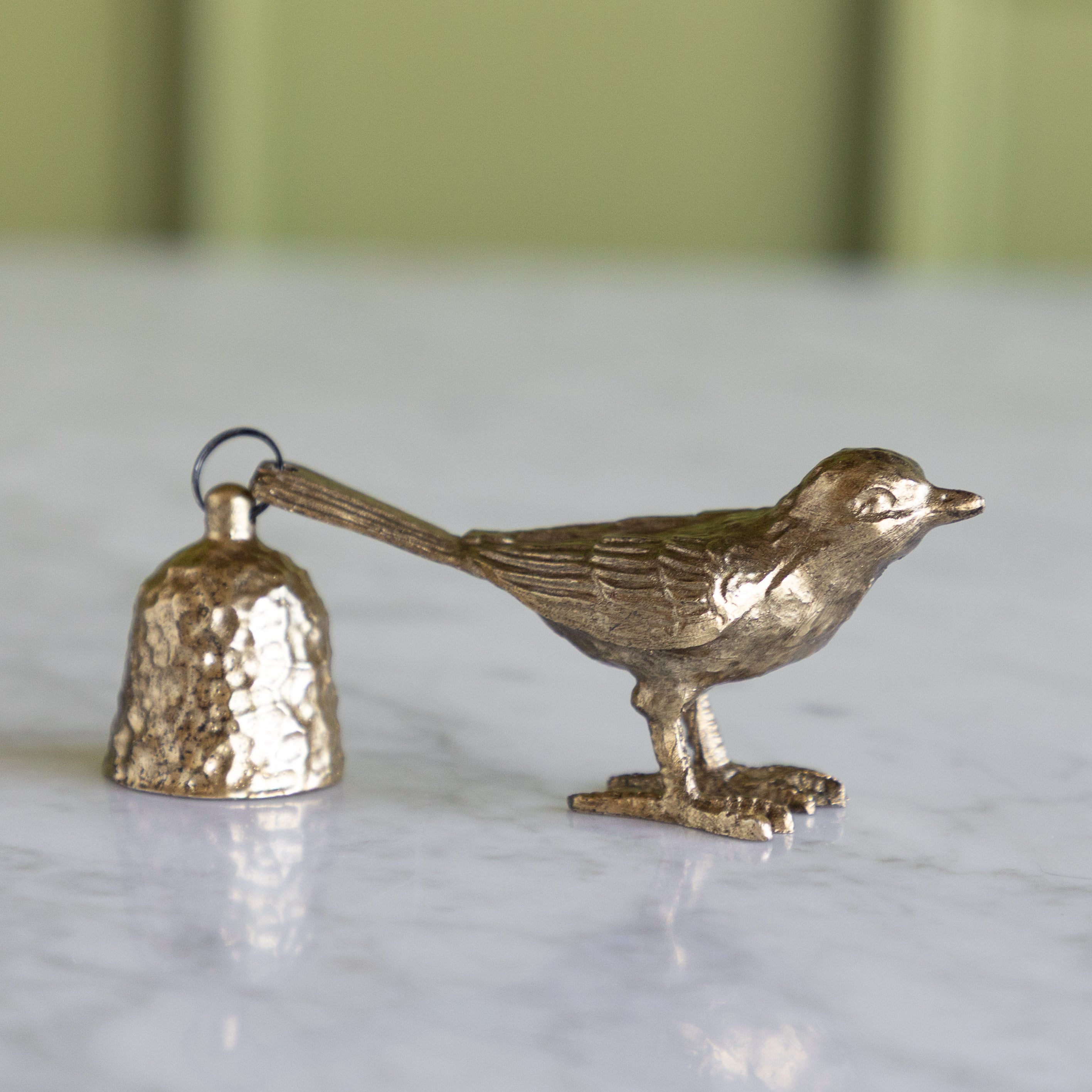 Bronze bird-shaped bell candle snuffer on a marble surface with a blurred green background.