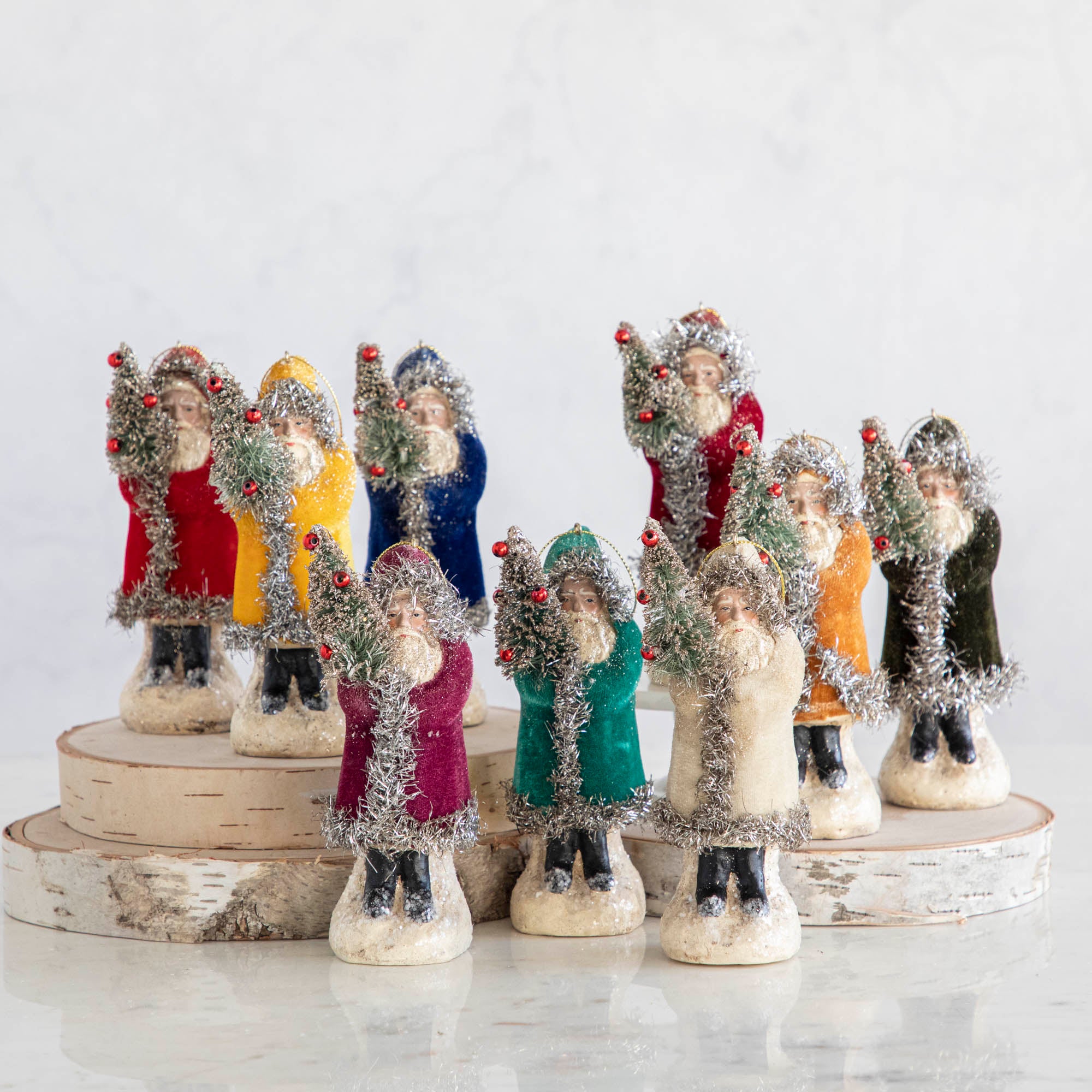 A collection of velvet-coated Santa ornaments in various colors, standing 5-6 inches tall, displayed on a wooden platform.