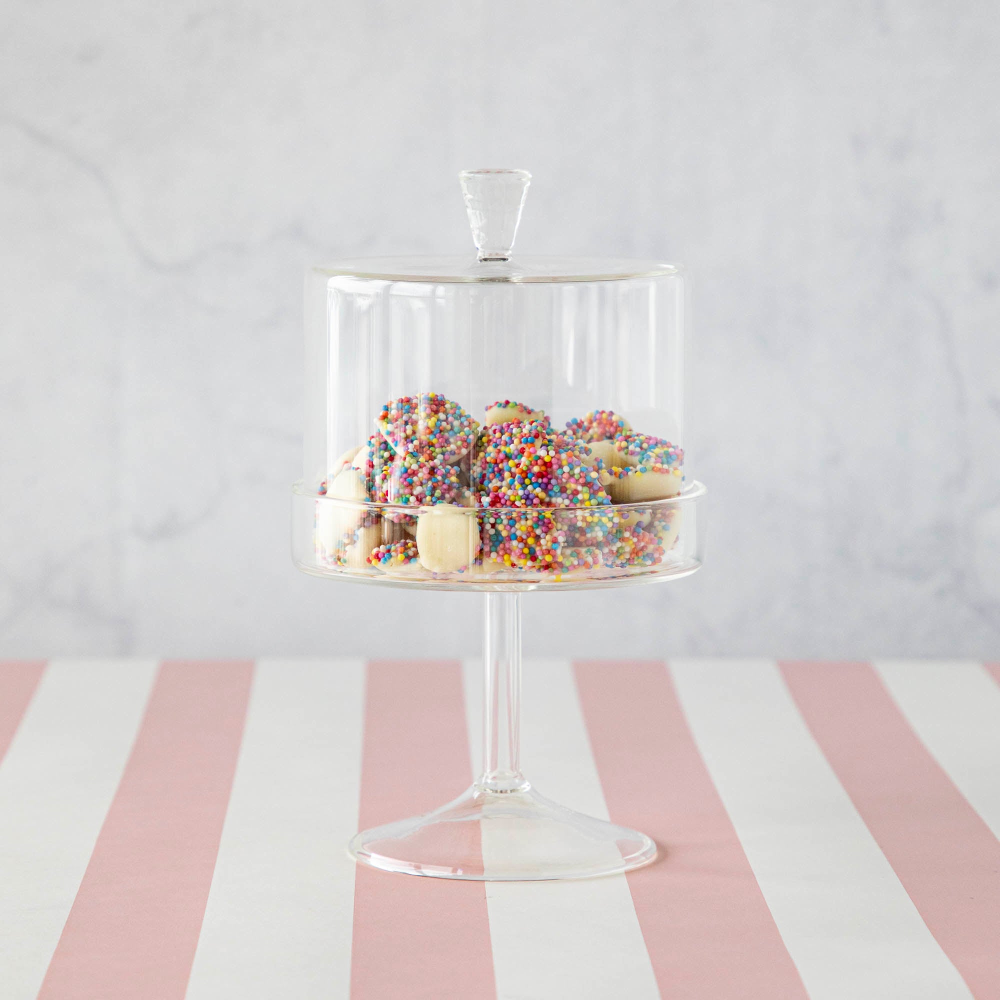 A HomArt Glass Dome & Pedestal cake stand with a glass dome lid filled with sprinkle-covered cookies on a pink and white striped surface.