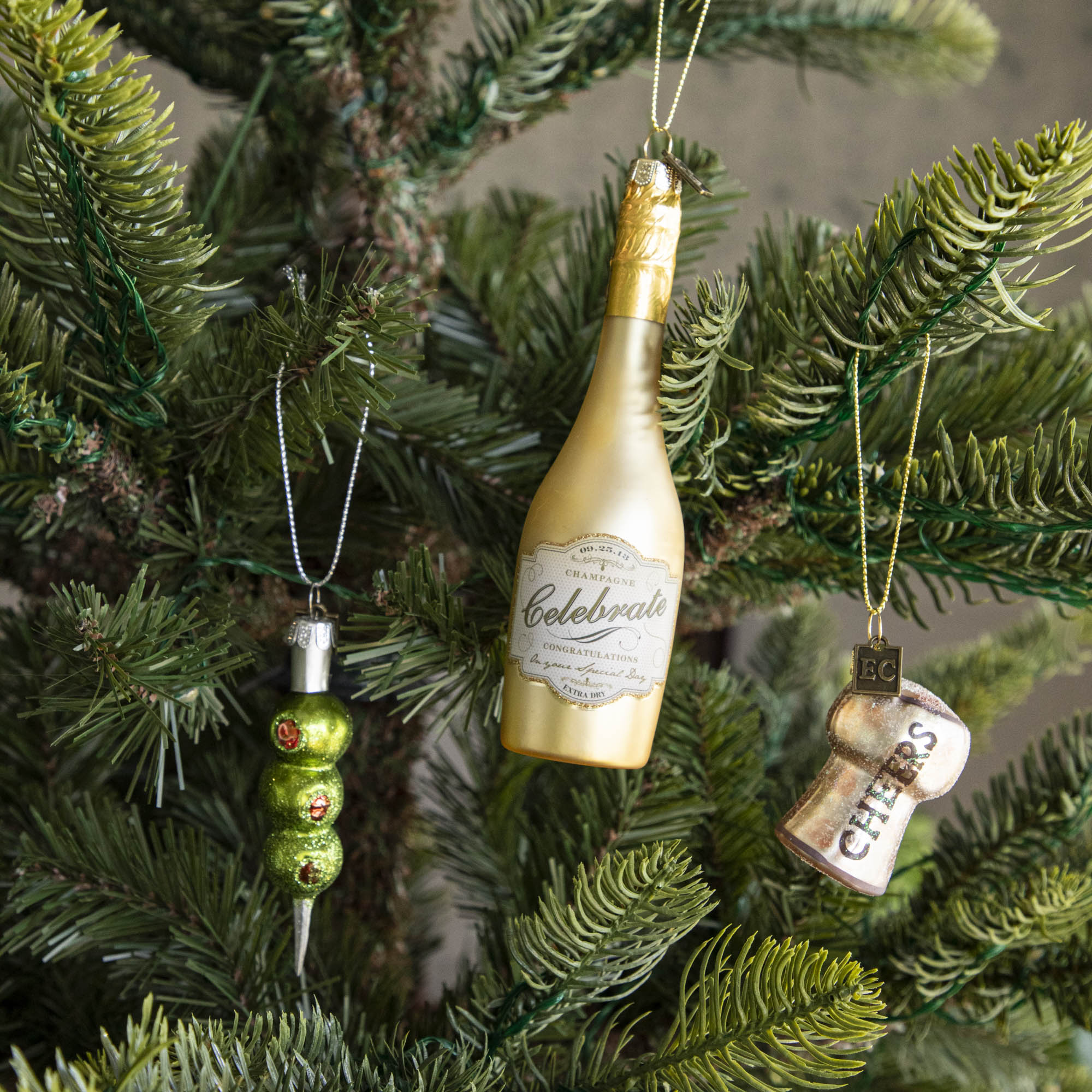 Three Olives Please Ornament, Champagne Ornament and Cheers Cork Ornament hanging on a tree.
