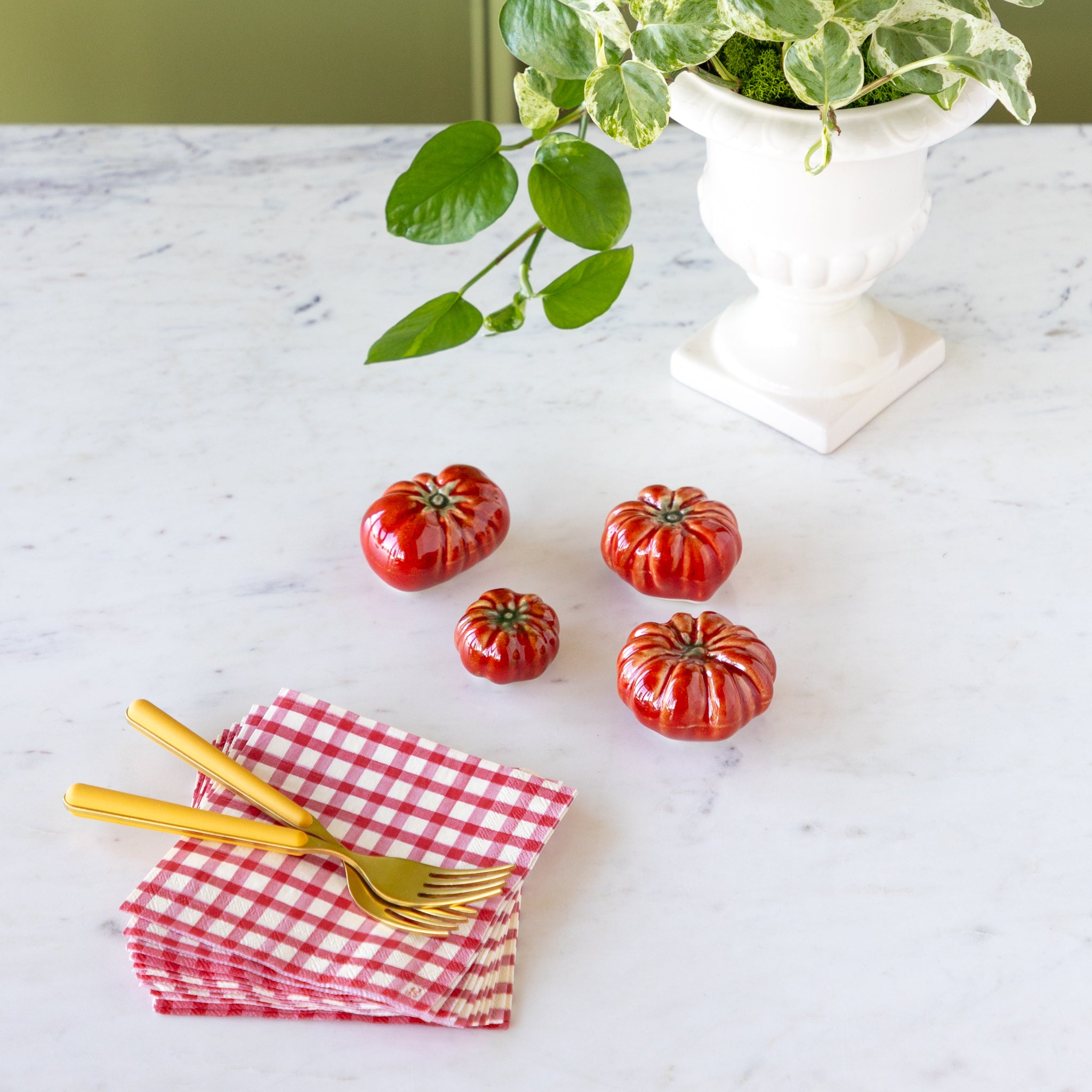 Red ceramic tomatoes on a marble surface with gold forks and a red checkered napkin.