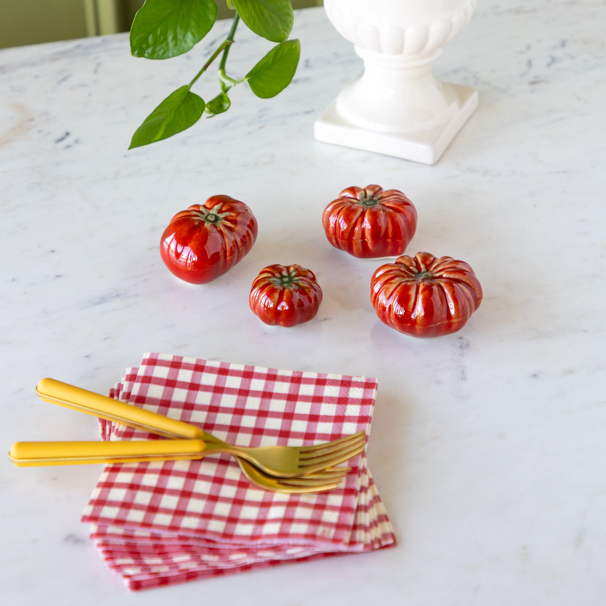 Red ceramic tomatoes on a marble surface with gold forks and a red checkered napkin.