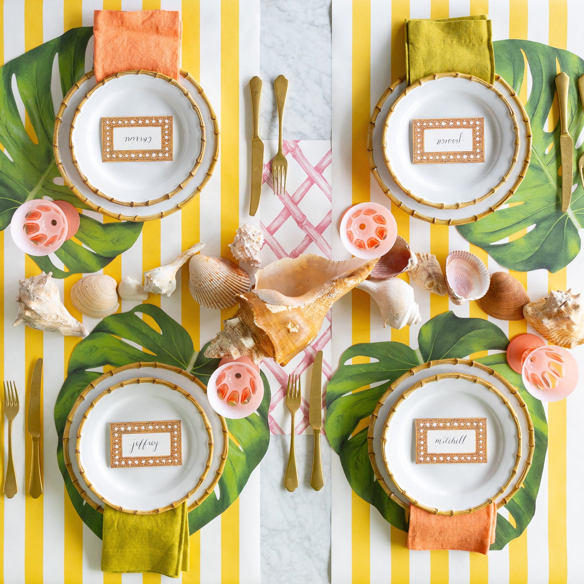 A tropical table setting featuring the Die-cut Monstera Leaf Placemats, bamboo rimmed dinner plates with the Rattan Weave Place Cards atop, seashells, gold flatware and green and peach napkins on the Marigold and Pink Lattice Runners.