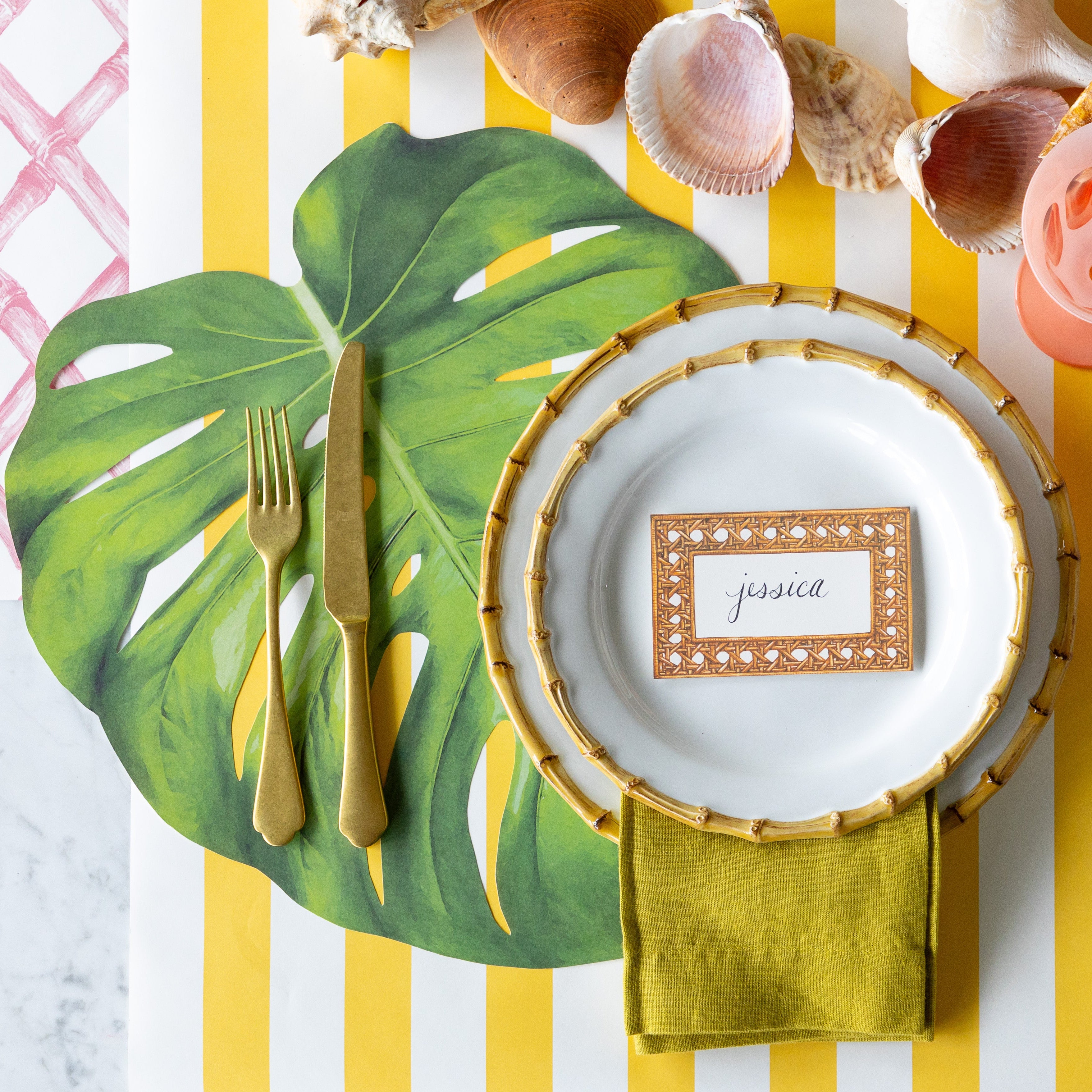 A tropical place setting featuring the Die-cut Monstera Leaf Placemat, bamboo rimmed dinner plates with the Rattan Weave Place Card reading "jessica" atop, seashells, gold flatware and a green napkin on the Marigold and Pink Lattice Runners.