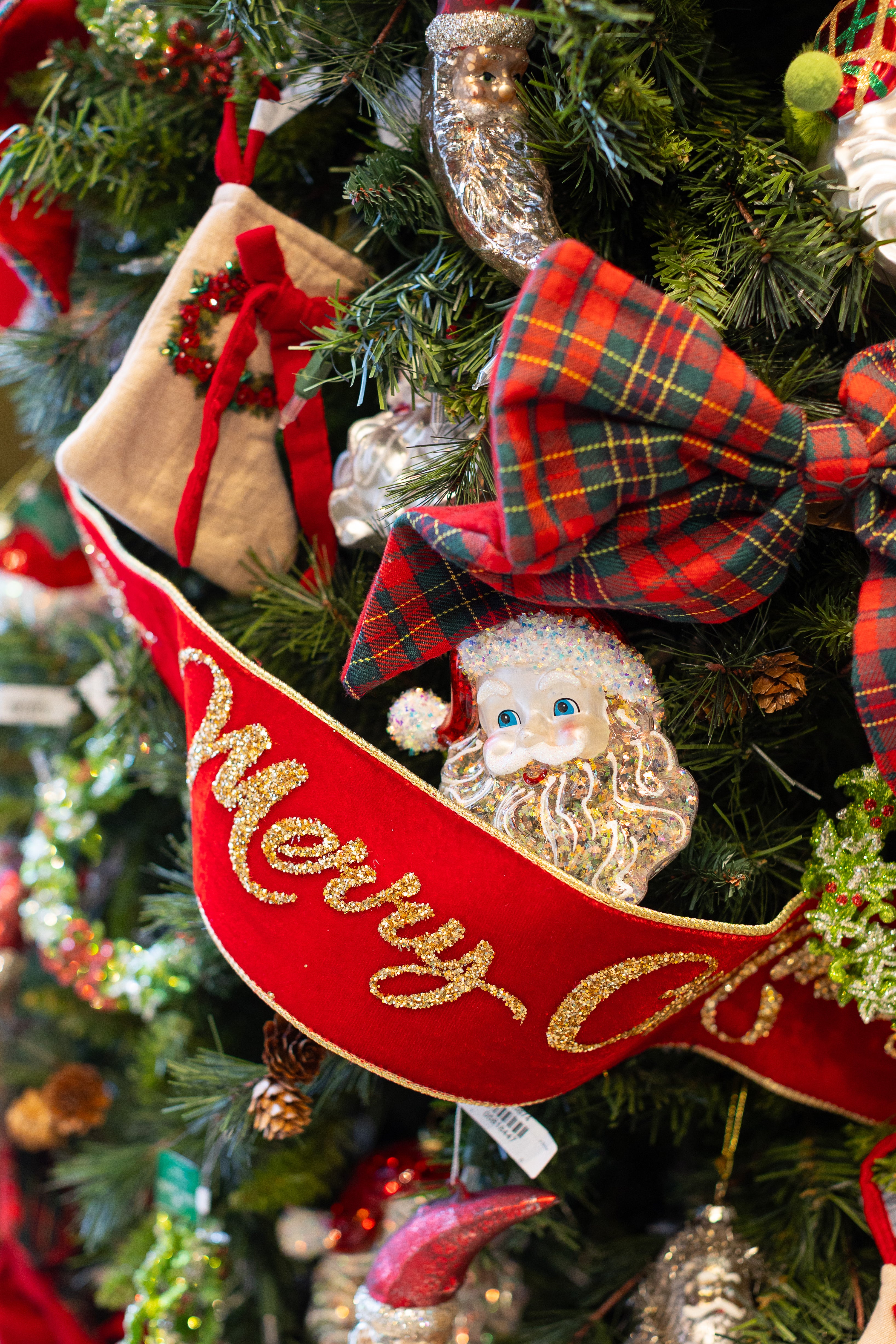Red ribbon with gold Merry Christmas written on it hanging on a Christmas tree with festive ornaments.
