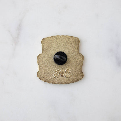 A Hester &amp; Cook Birthday Cake Enamel Pin on a marble surface.