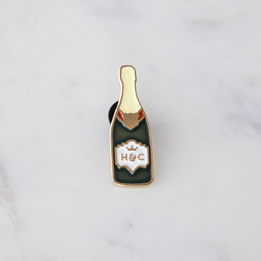 A gilded story envelops a bottle of Hester &amp; Cook Champagne Enamel Pin on a marble table.