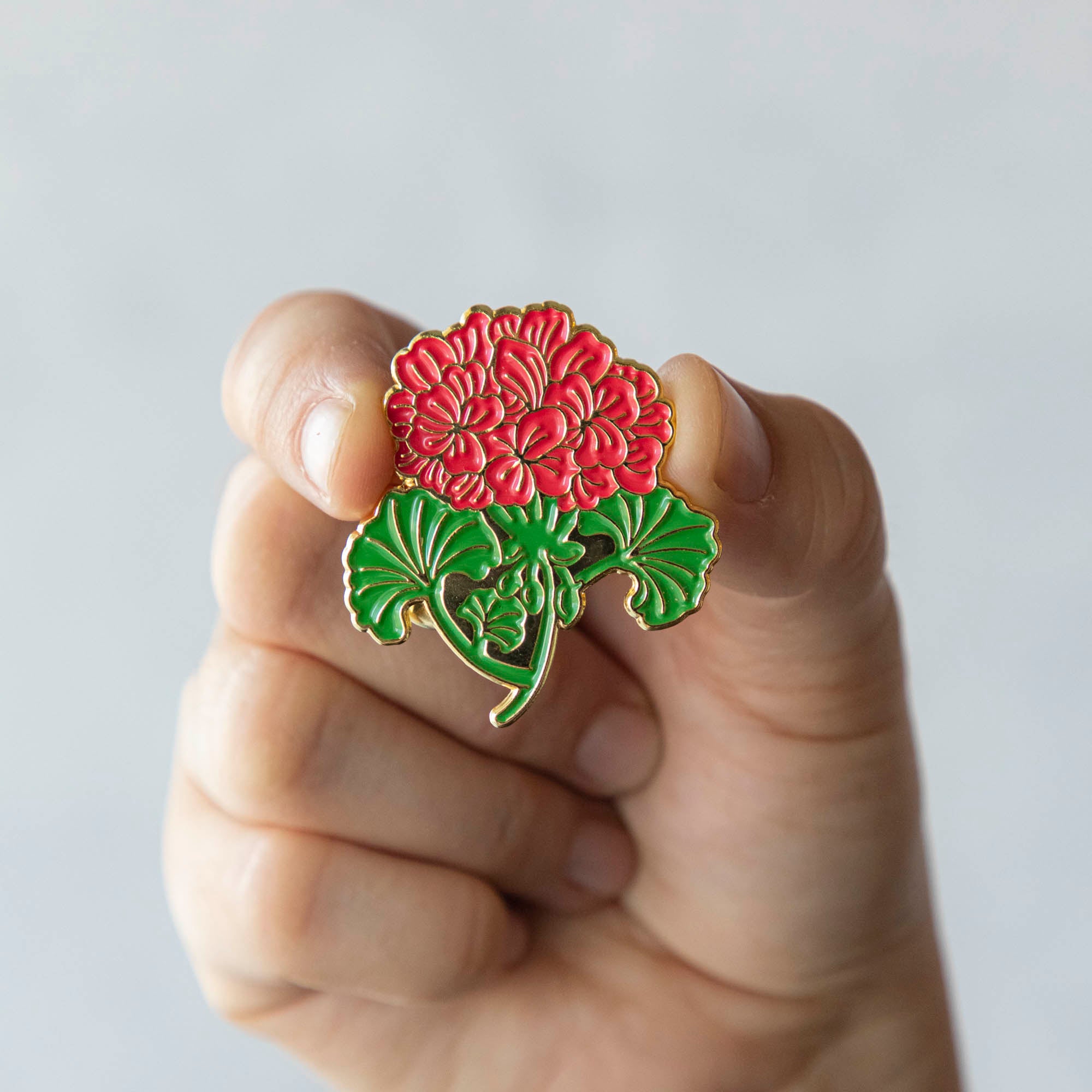A person proudly displaying a Hester & Cook Geranium Enamel Pin with a vibrant red chrysanthemum flower.